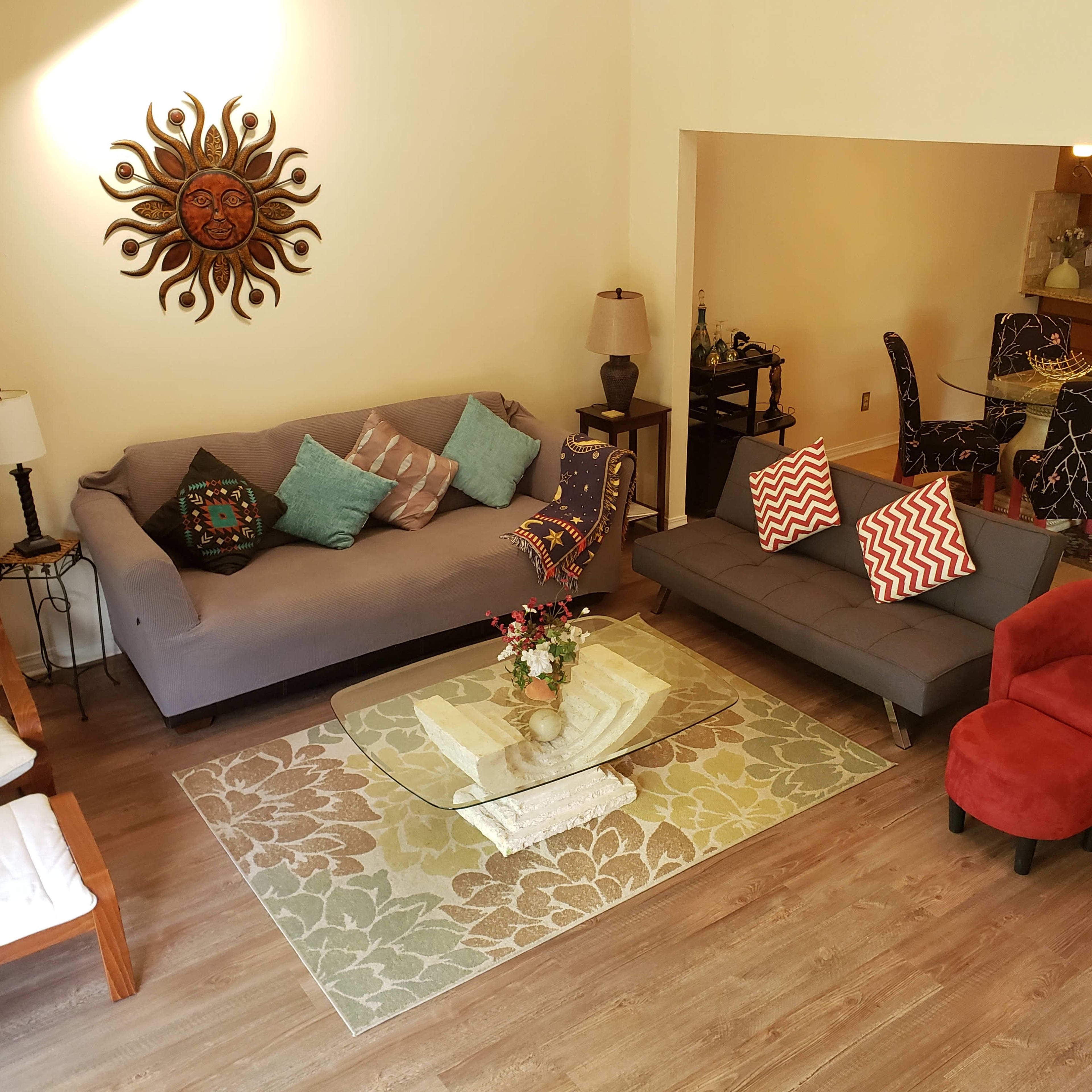 A living room area with two sofas, a glass coffee table, a decorative rug, and a wall-mounted sun sculpture.