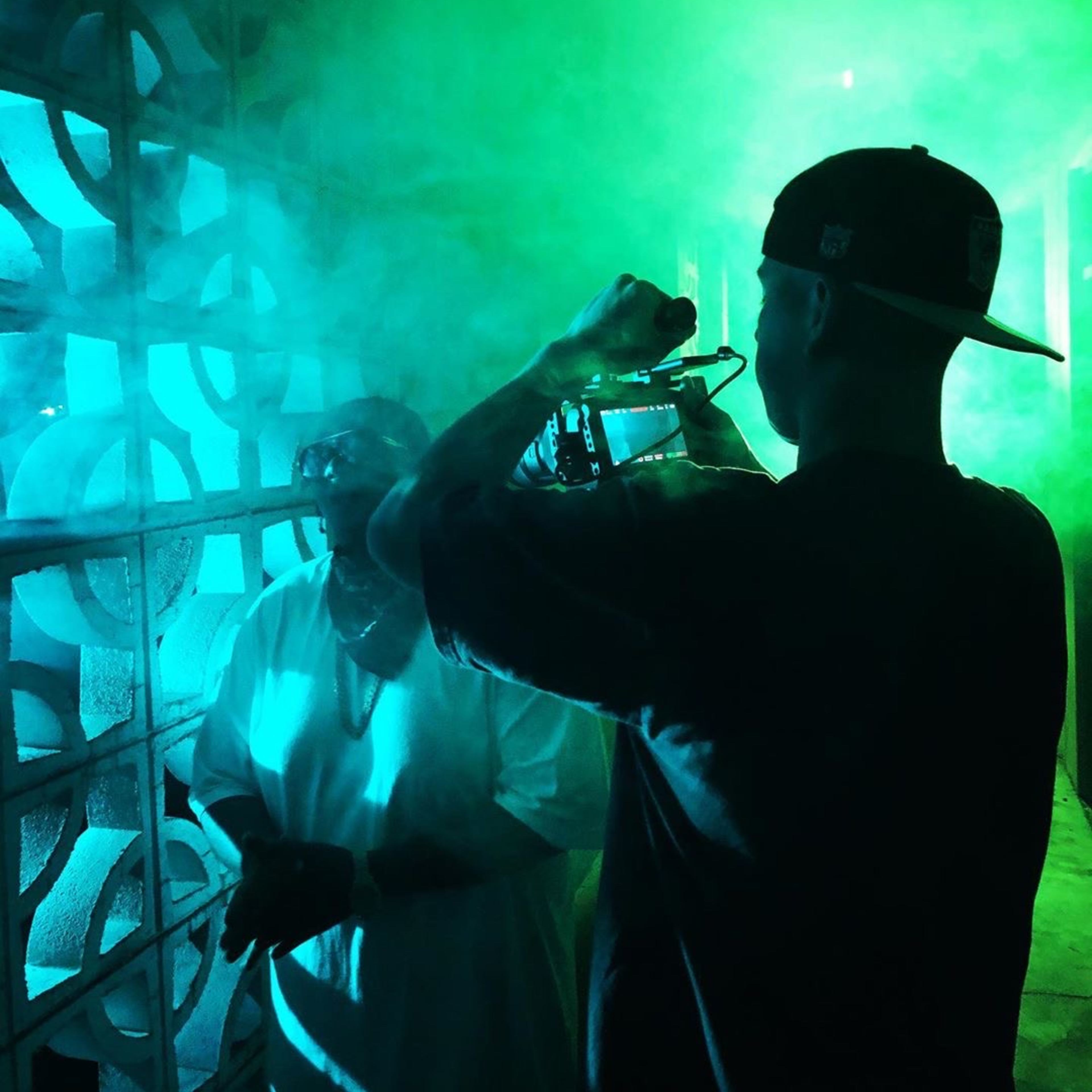A person holds a camera while another person stands in a foggy, illuminated setting with green and blue lights.