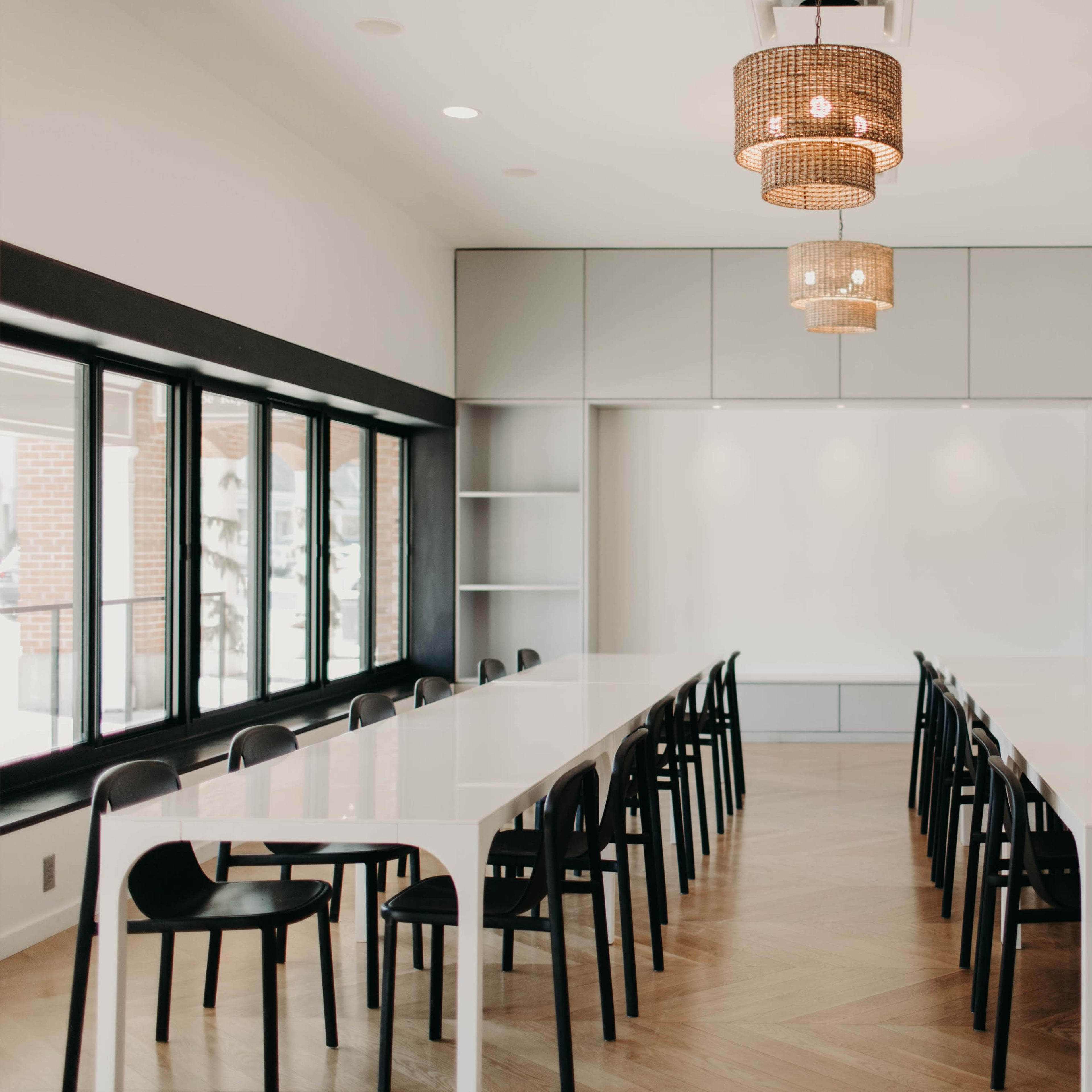 A modern conference room features a long white table surrounded by black chairs, illuminated by stylish pendant lights.