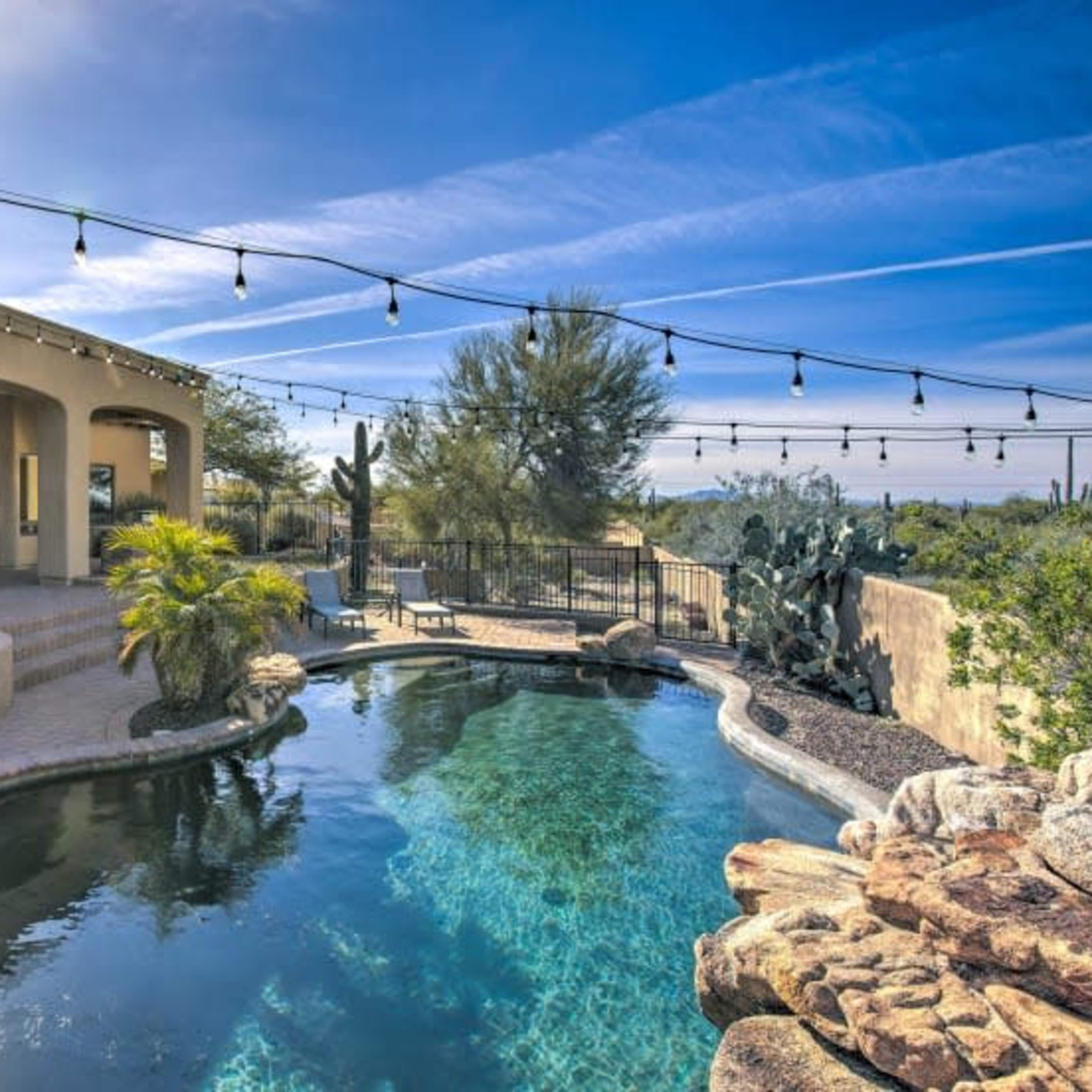 A backyard pool area with a hot tub, surrounded by desert landscaping and strung lights under a clear blue sky.