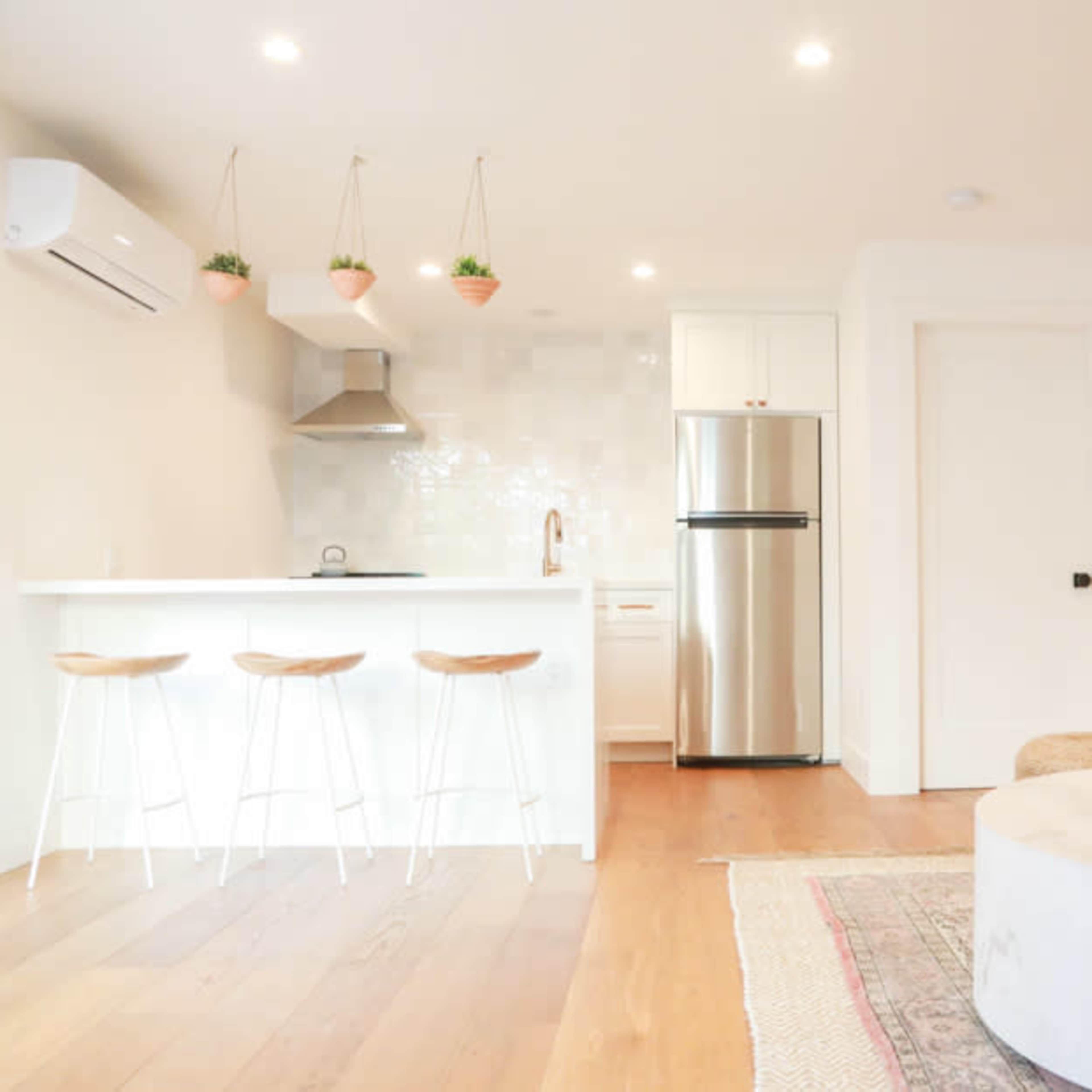 A modern kitchen and living area with a white counter, stainless steel fridge, and decorative plants hanging above bar stools.