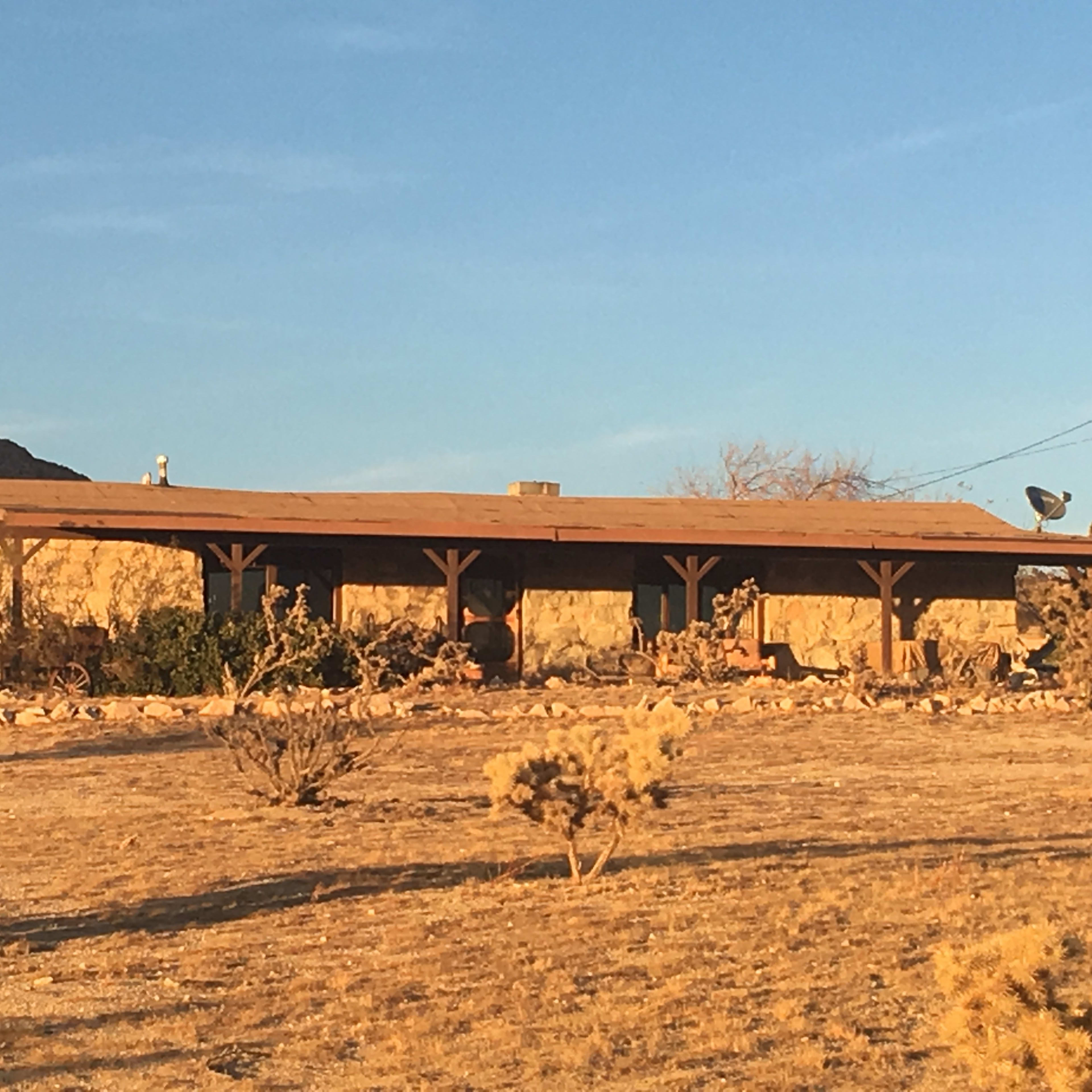 A single-story adobe house stands in a barren landscape with sparse vegetation and distant mountains.