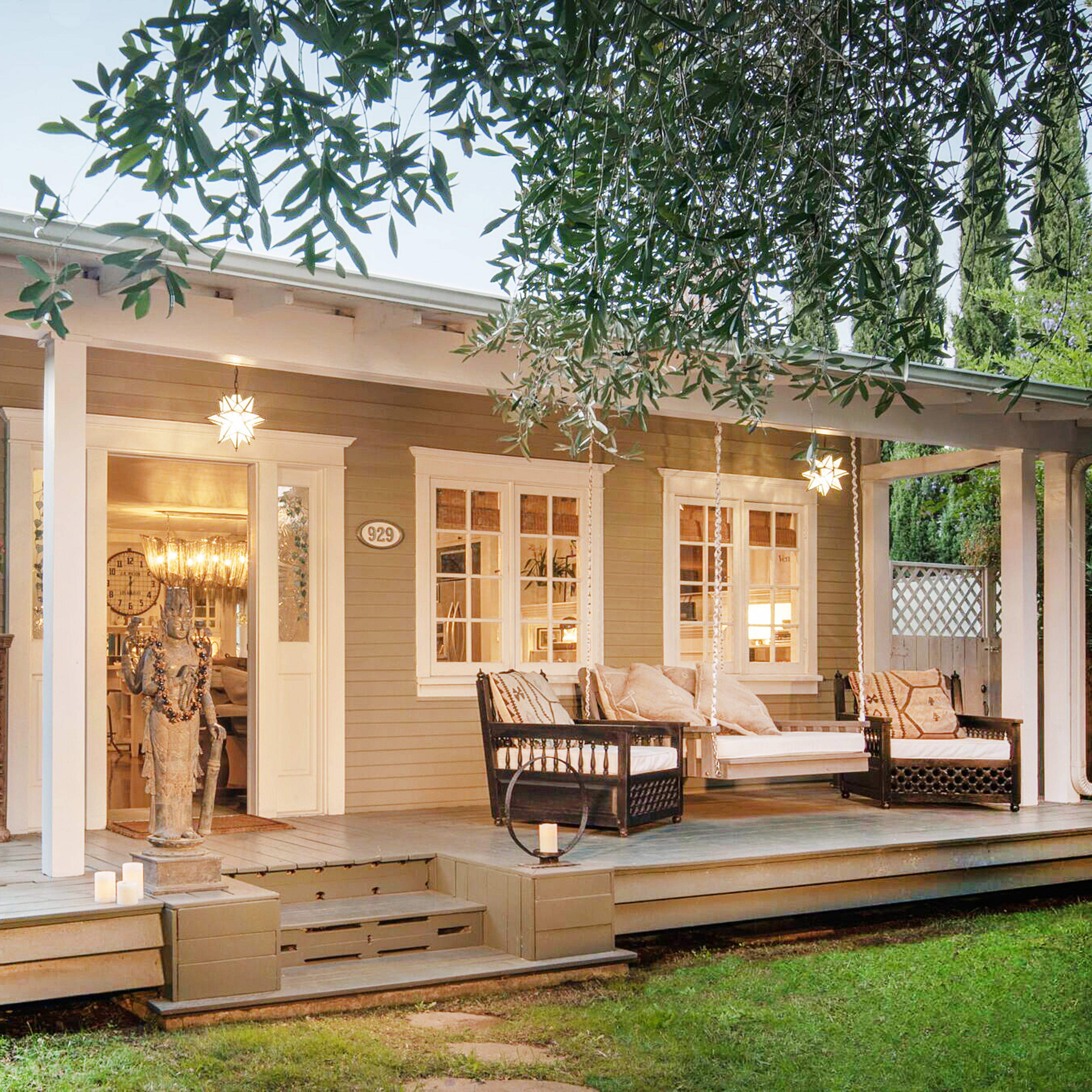 A cozy, light-colored house with a front porch, featuring comfortable seating, surrounded by greenery.
