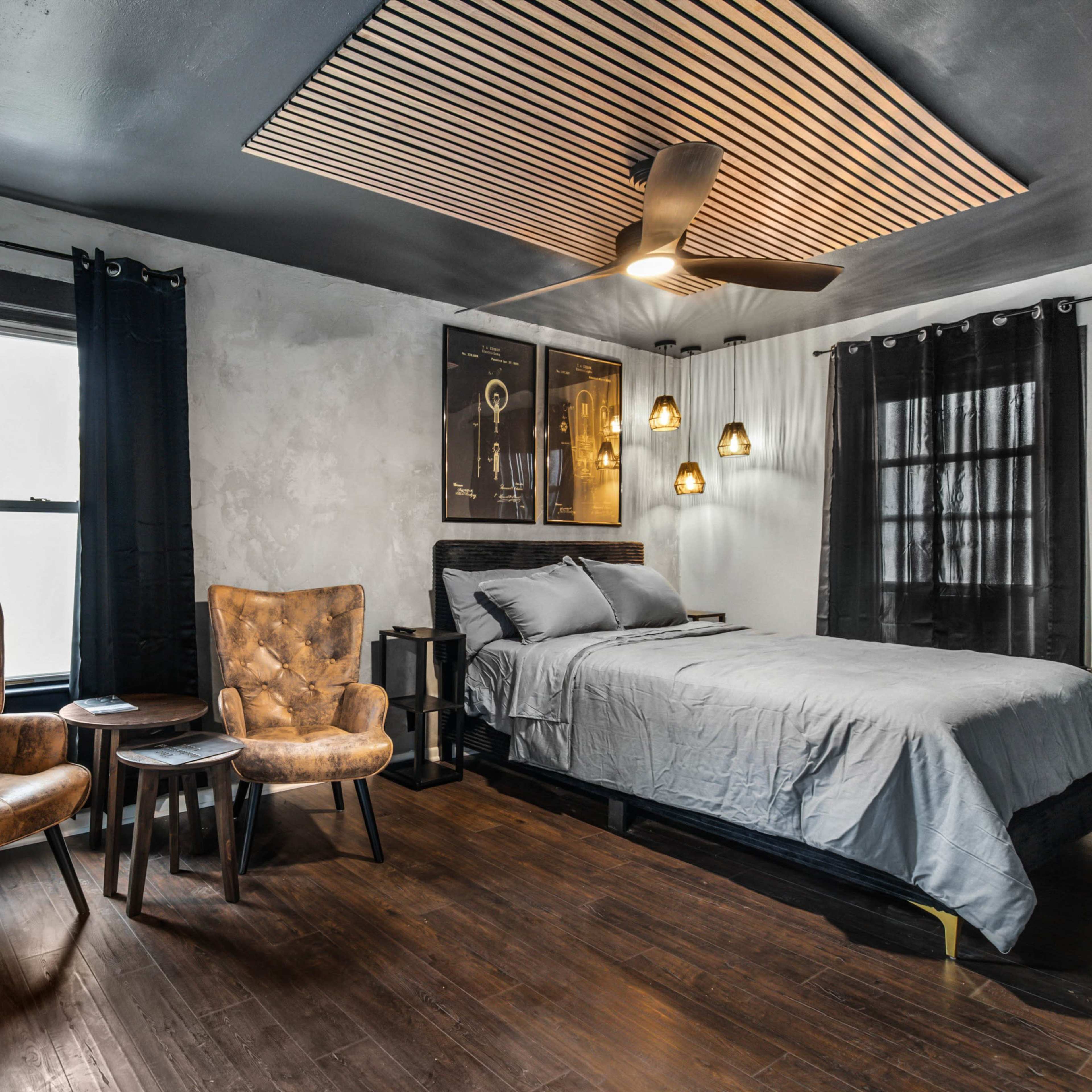 A bedroom featuring a bed, two armchairs, and a wooden ceiling design, with dark curtains and a wooden floor.