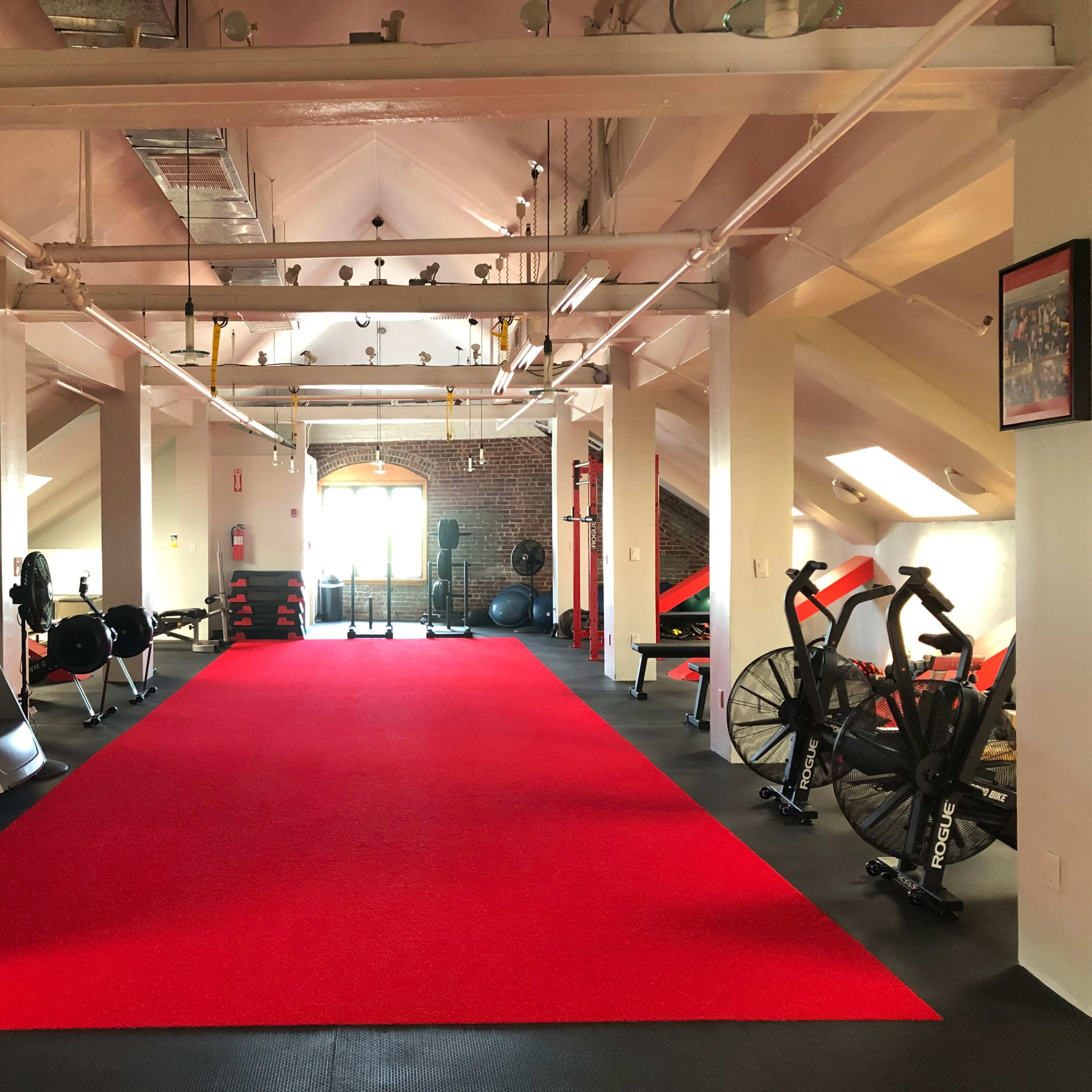 A well-lit gym with red carpet flooring, featuring several exercise machines, free weights, and workout equipment arranged along the walls.