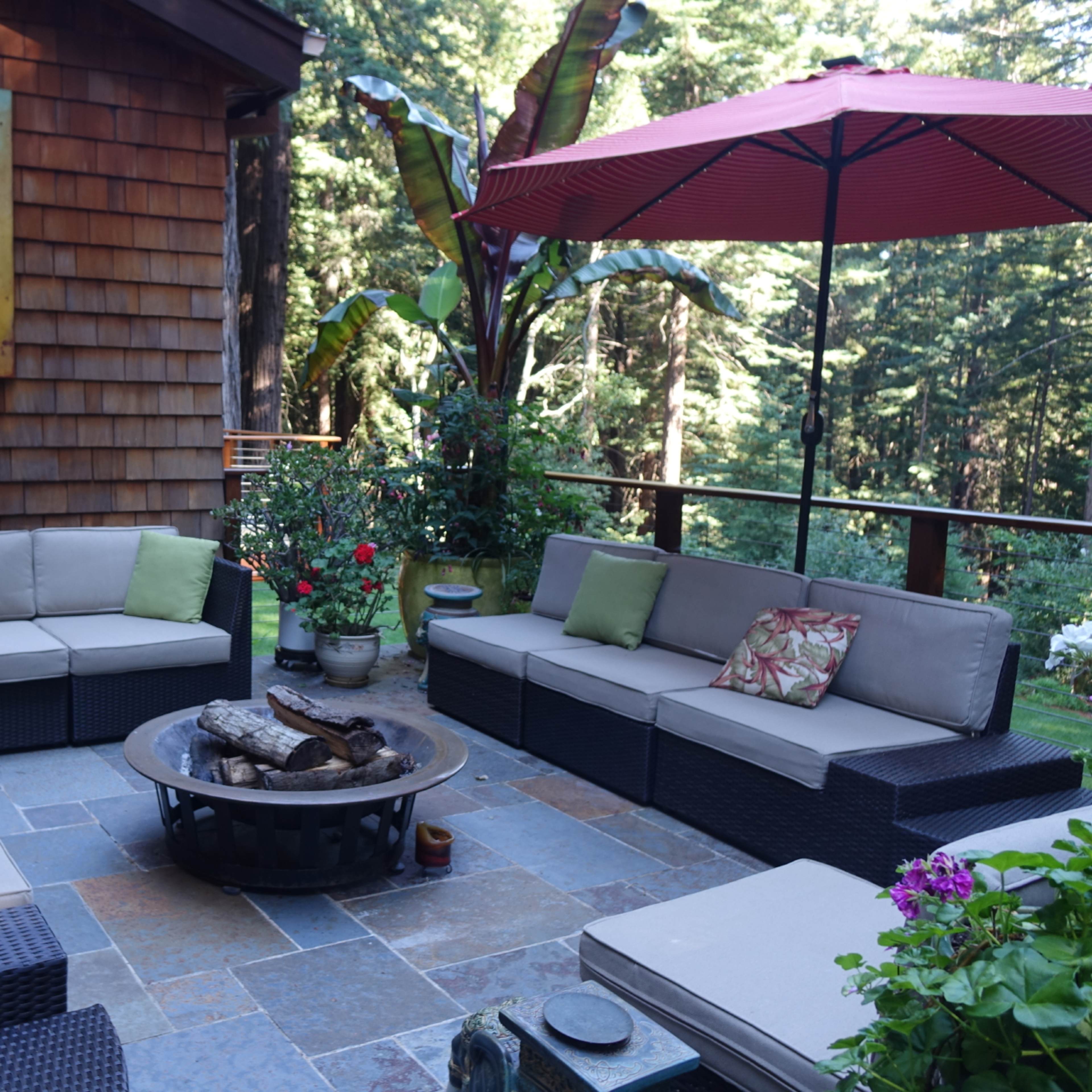 A patio area features sectional seating, a fire pit, and an umbrella, surrounded by greenery and a wooden house in the background.
