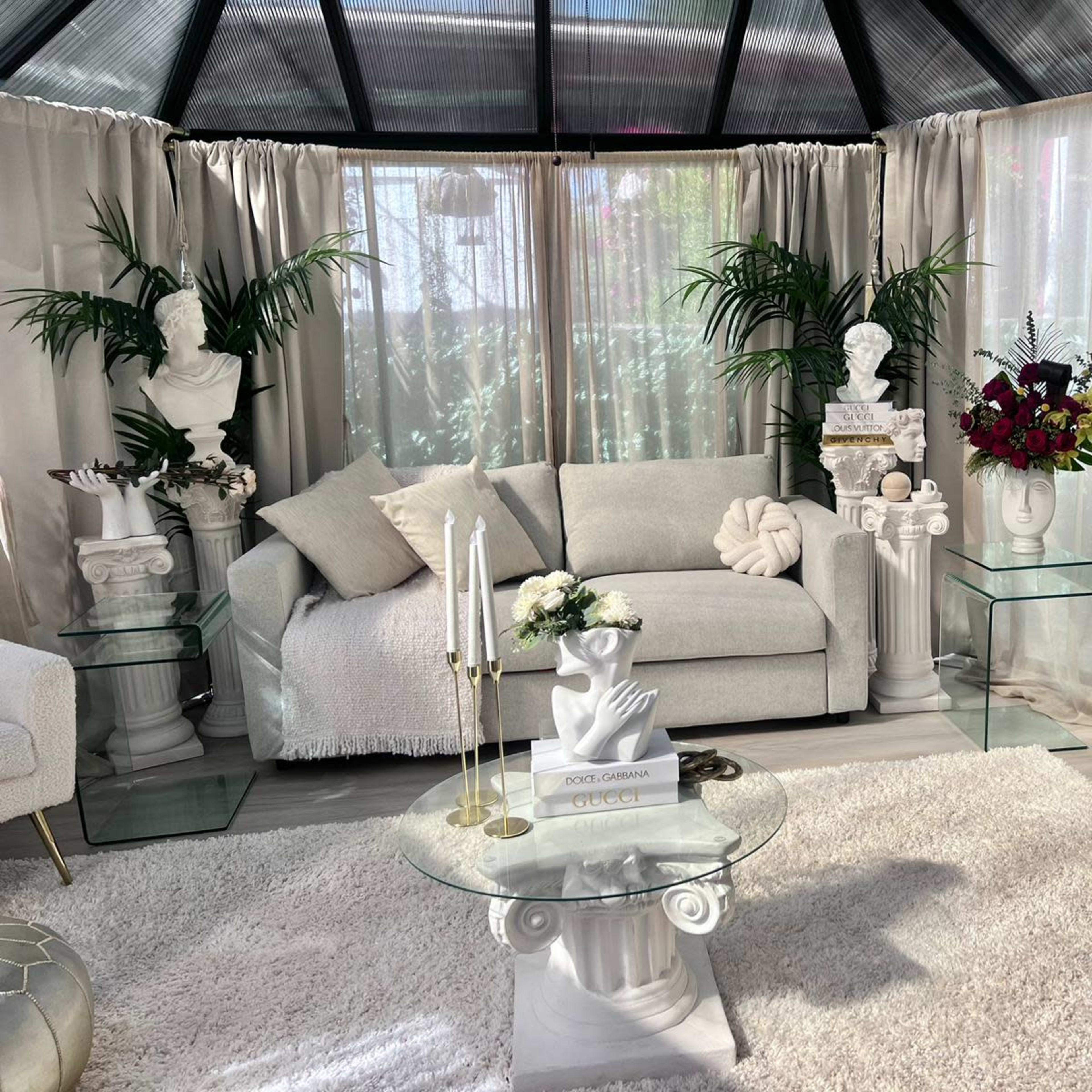 A modern indoor seating area featuring a light-colored sofa, glass tables, and decorative busts amidst greenery and floral arrangements.