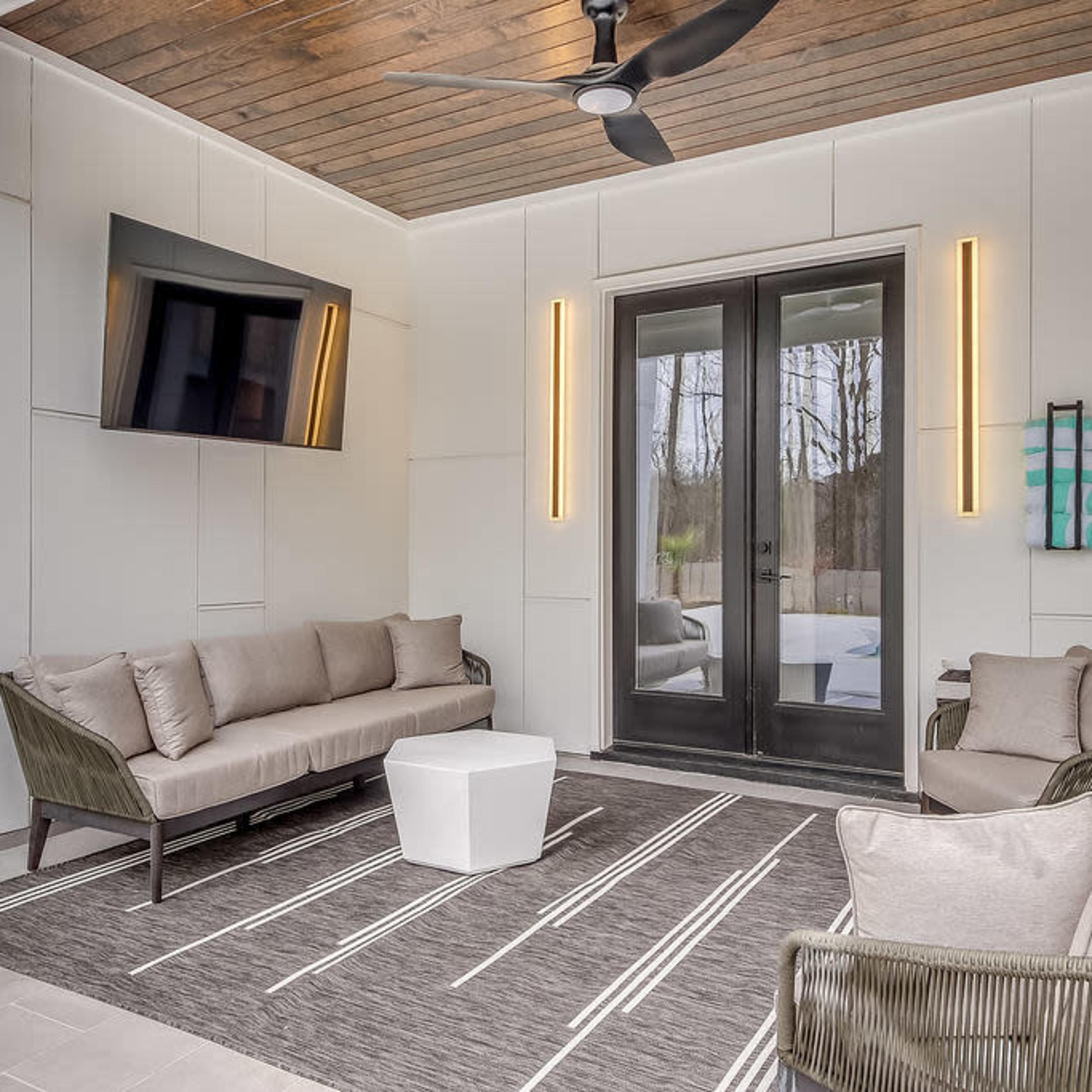 A modern, covered outdoor living space with two couches, a television mounted on the wall, and large glass doors leading to an outdoor area.