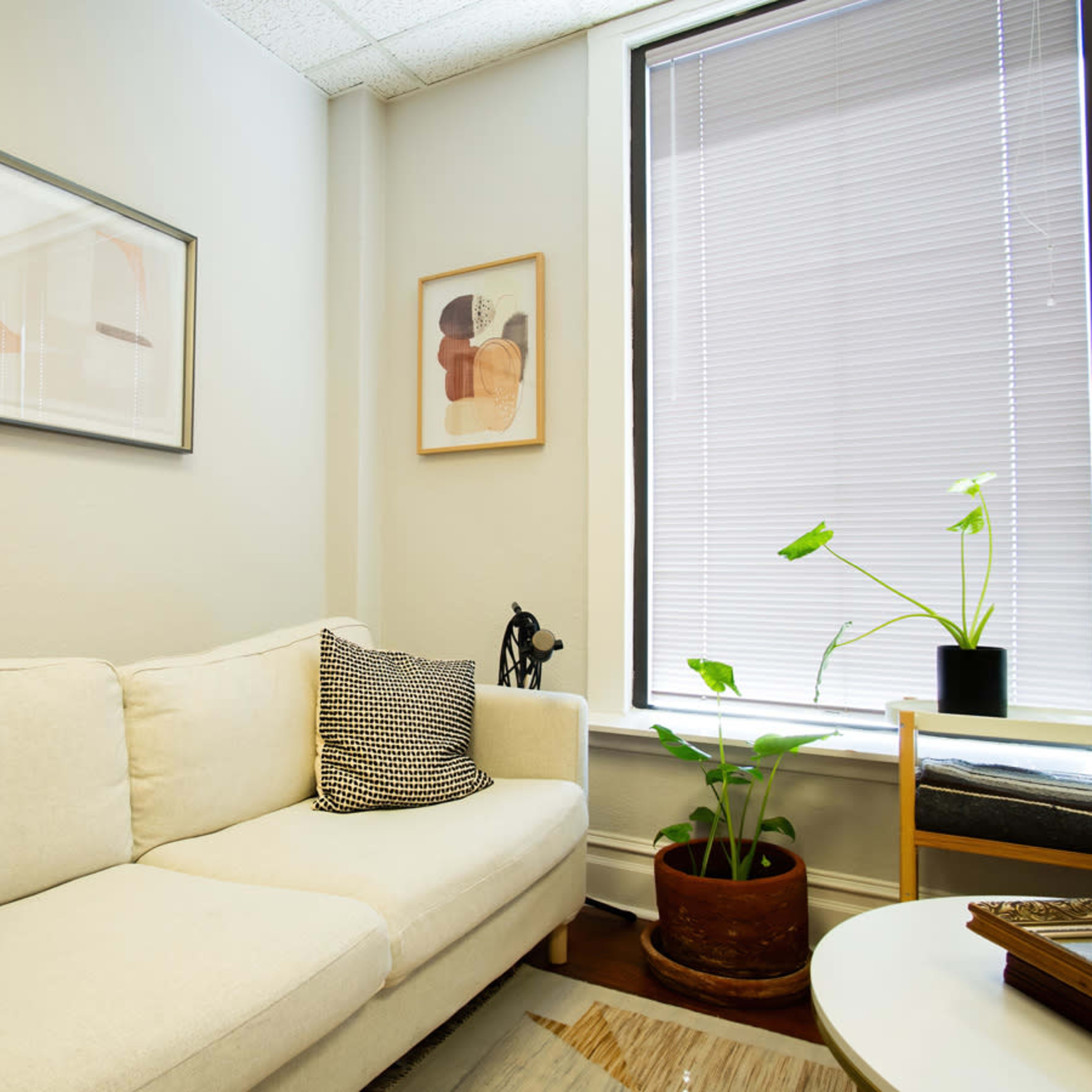 A light-colored sofa with two patterned pillows is positioned against a wall, accompanied by framed artwork and a potted plant in a window nook.