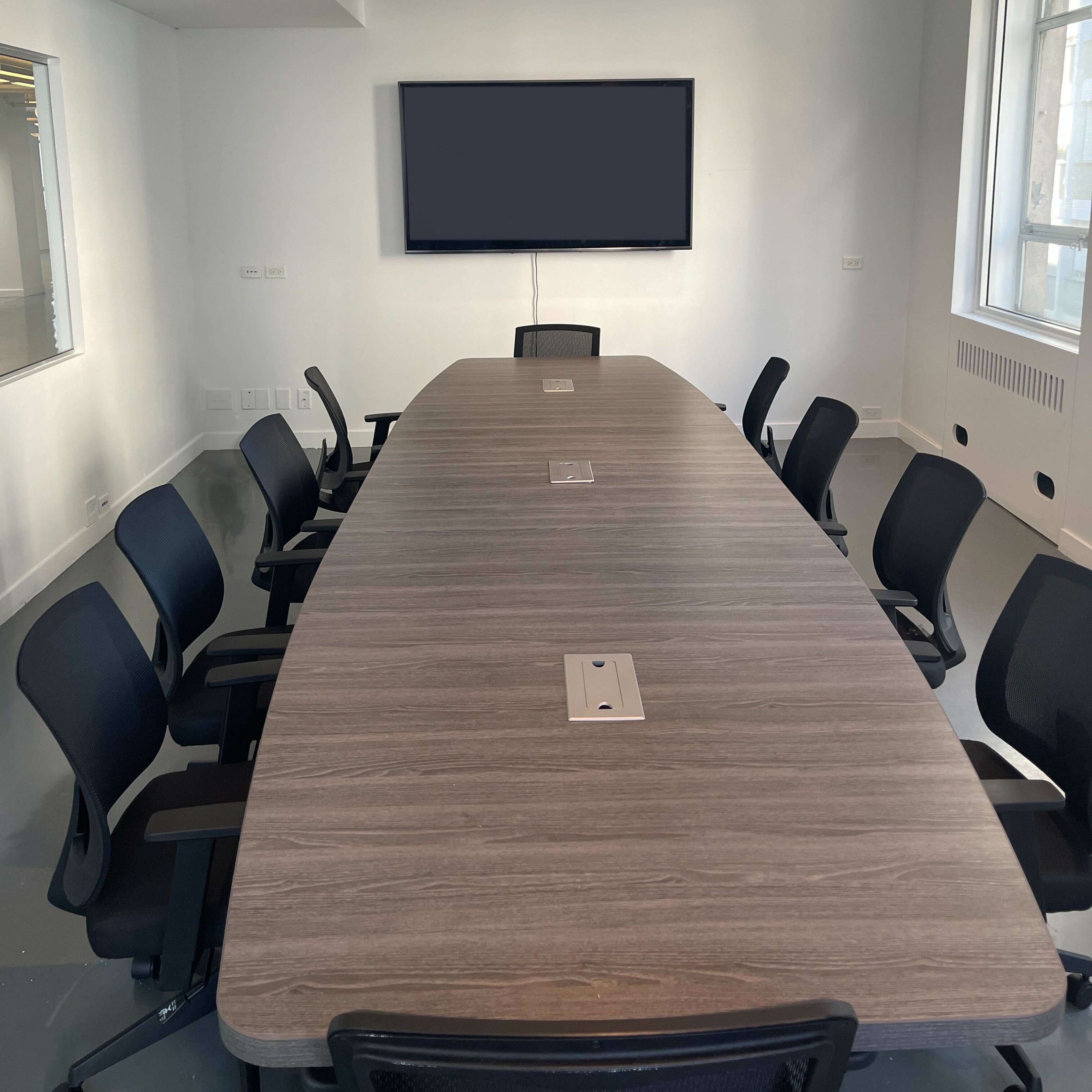 A large, oval conference table is surrounded by black ergonomic chairs in a brightly lit meeting room, featuring a wall-mounted screen and windows.