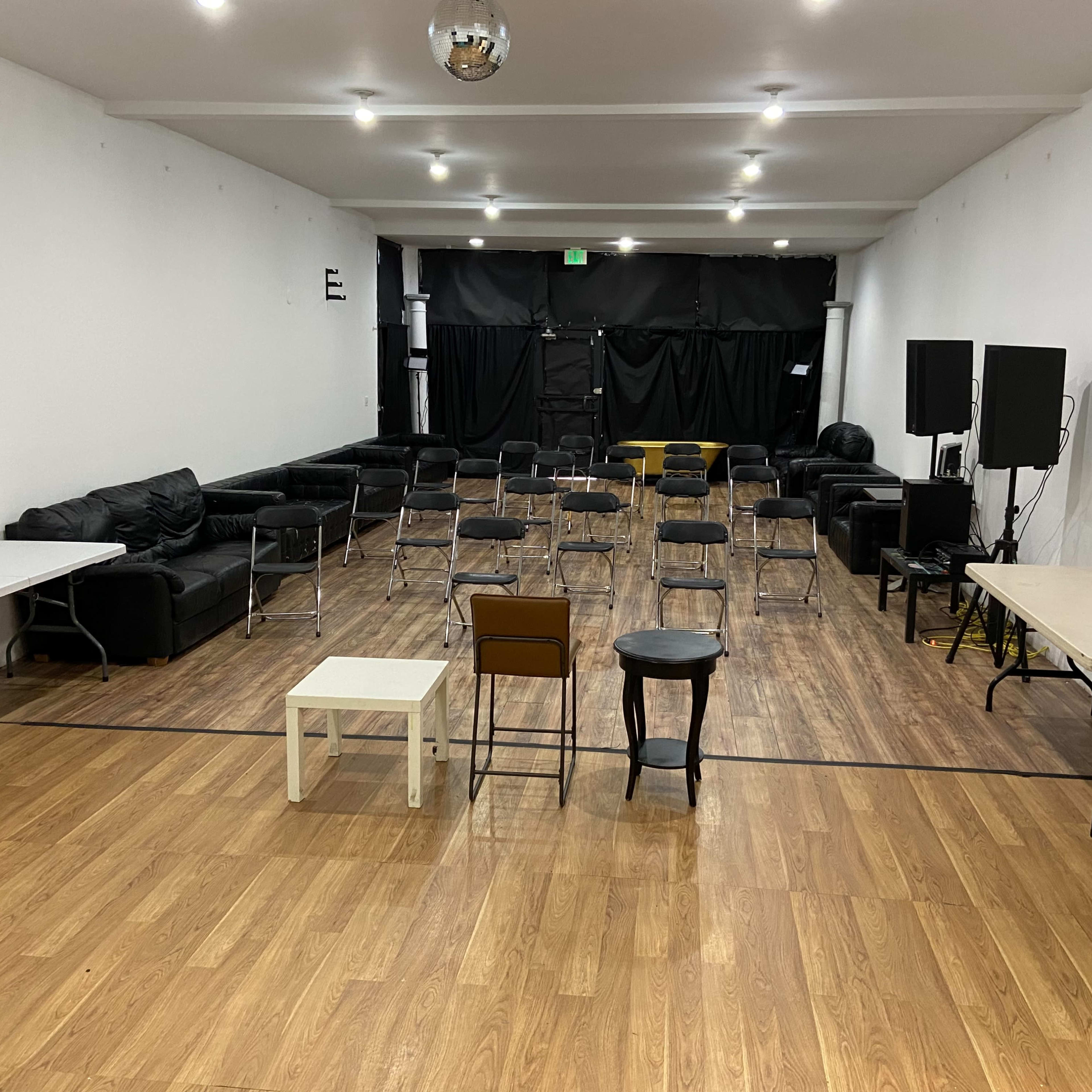 A spacious room with arranged chairs, tables, and black couches against white walls, illuminated by overhead lights.