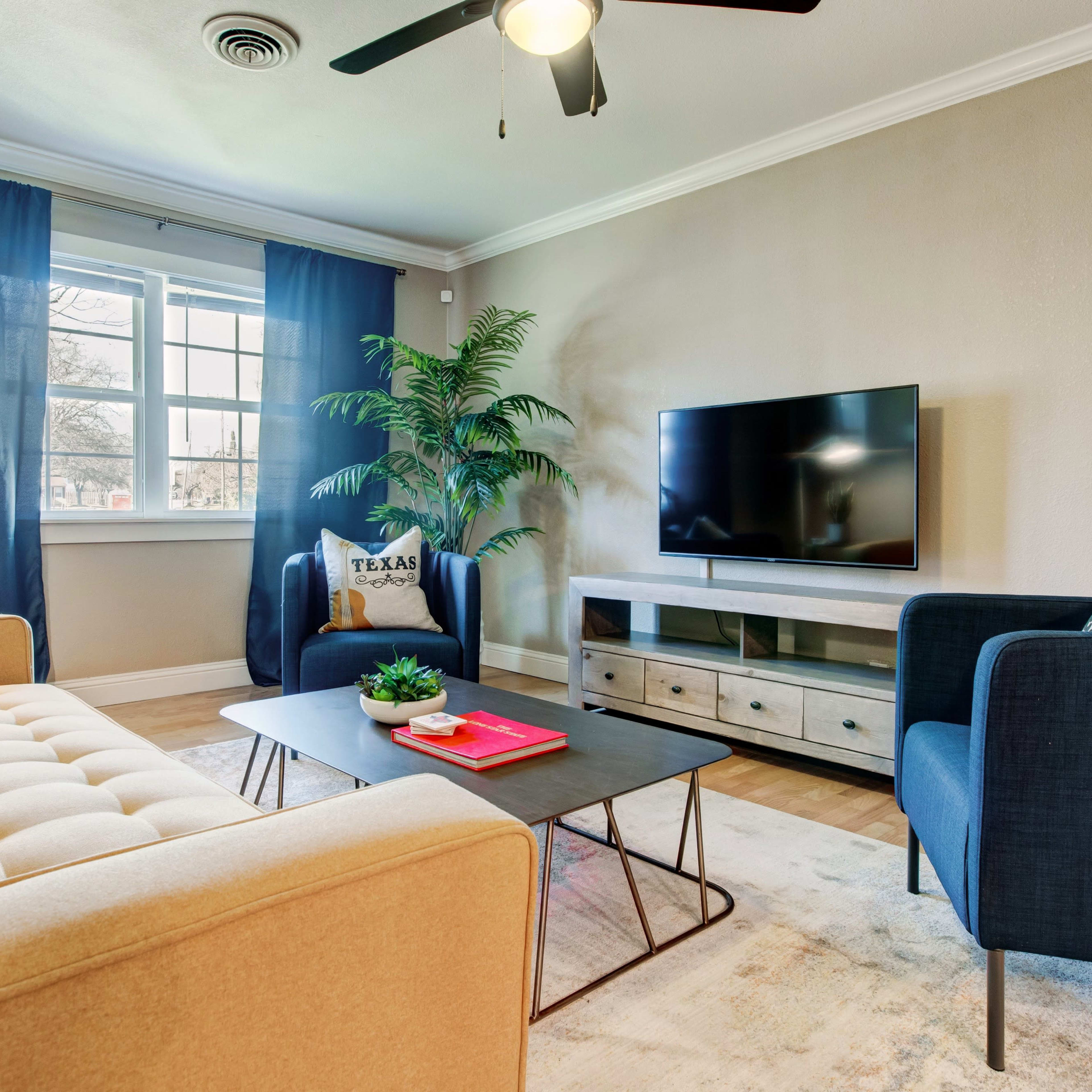 A living room with a beige sofa, a coffee table with books, a flat-screen TV on a cabinet, and potted plants against the backdrop of blue curtains.