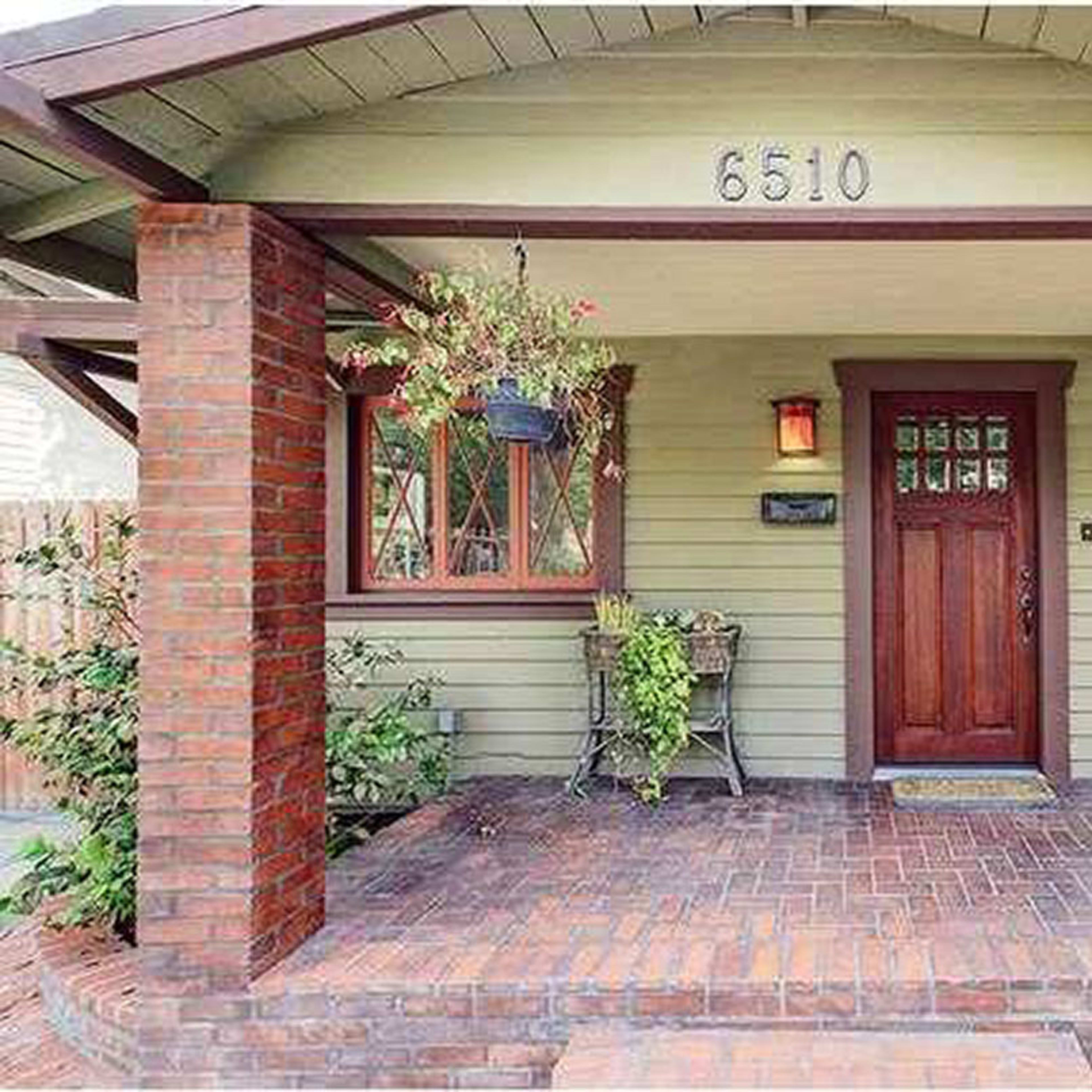 A front porch of a green bungalow with red brick pillars and a wooden door, featuring potted plants and a welcome mat.