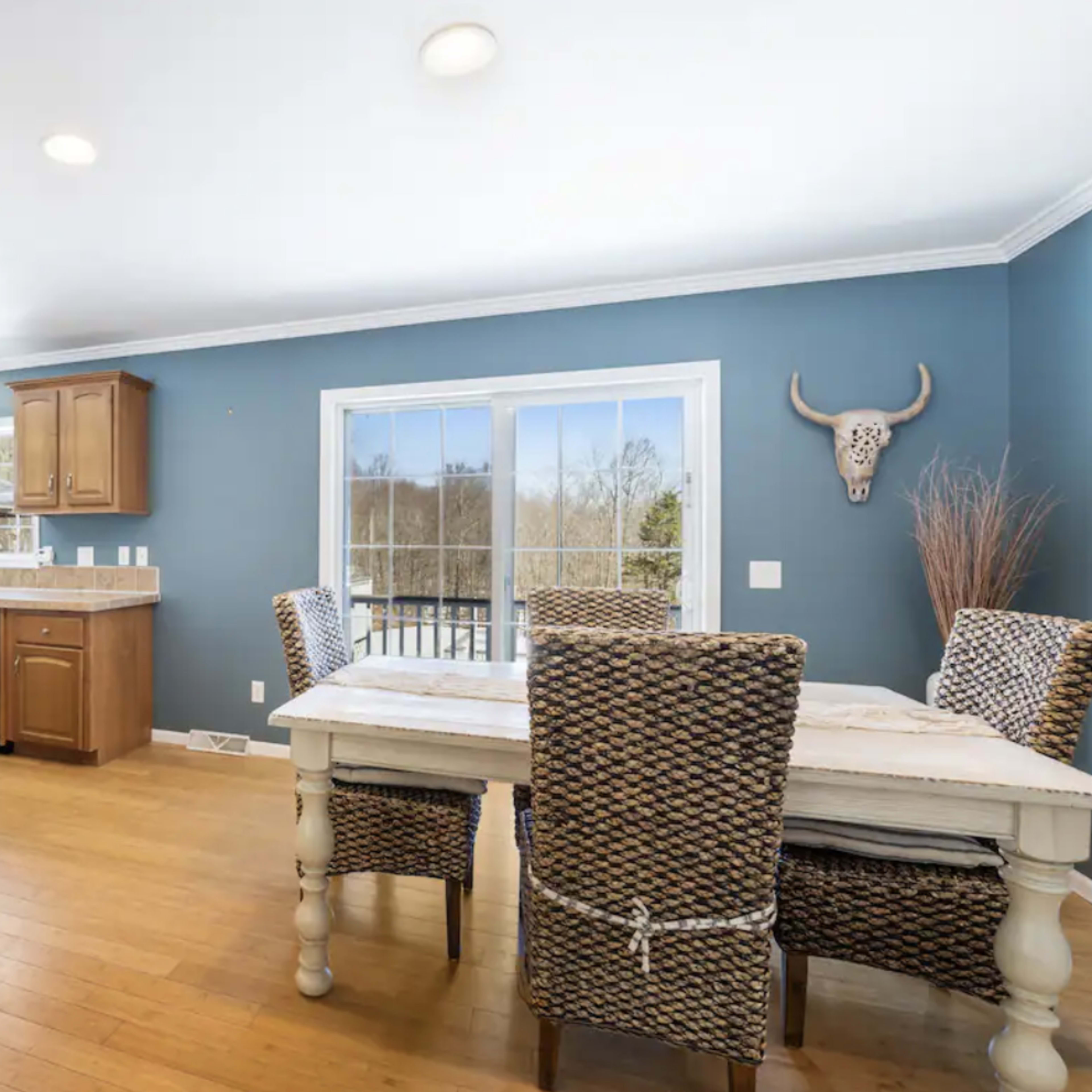 A kitchen area with wooden cabinets, a dining table with woven chairs, and large windows overlooking a natural landscape.