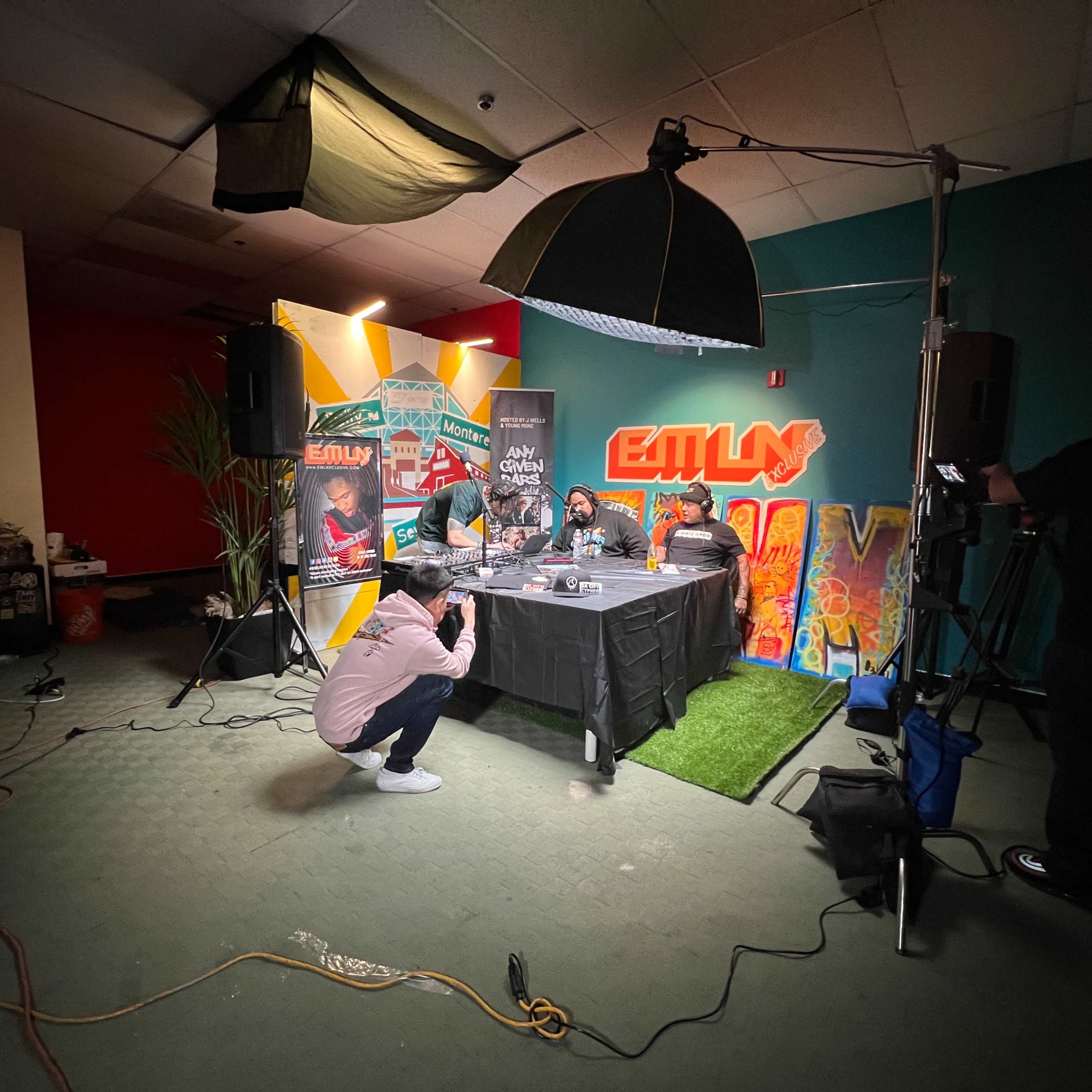 A group of people is seated at a table for a podcast recording, surrounded by colorful banners and lighting equipment in a commercial space.