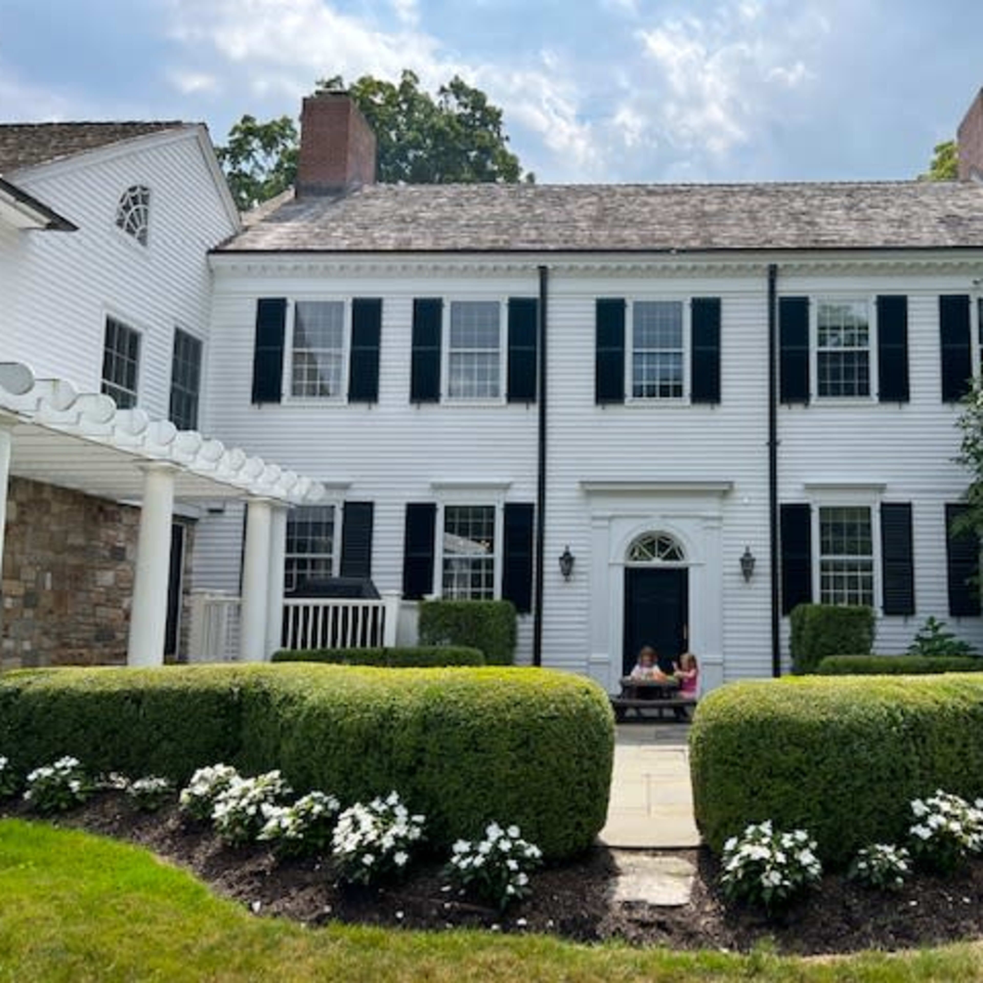 A large white colonial-style house with black shutters is surrounded by meticulously trimmed hedges and flower beds in front.