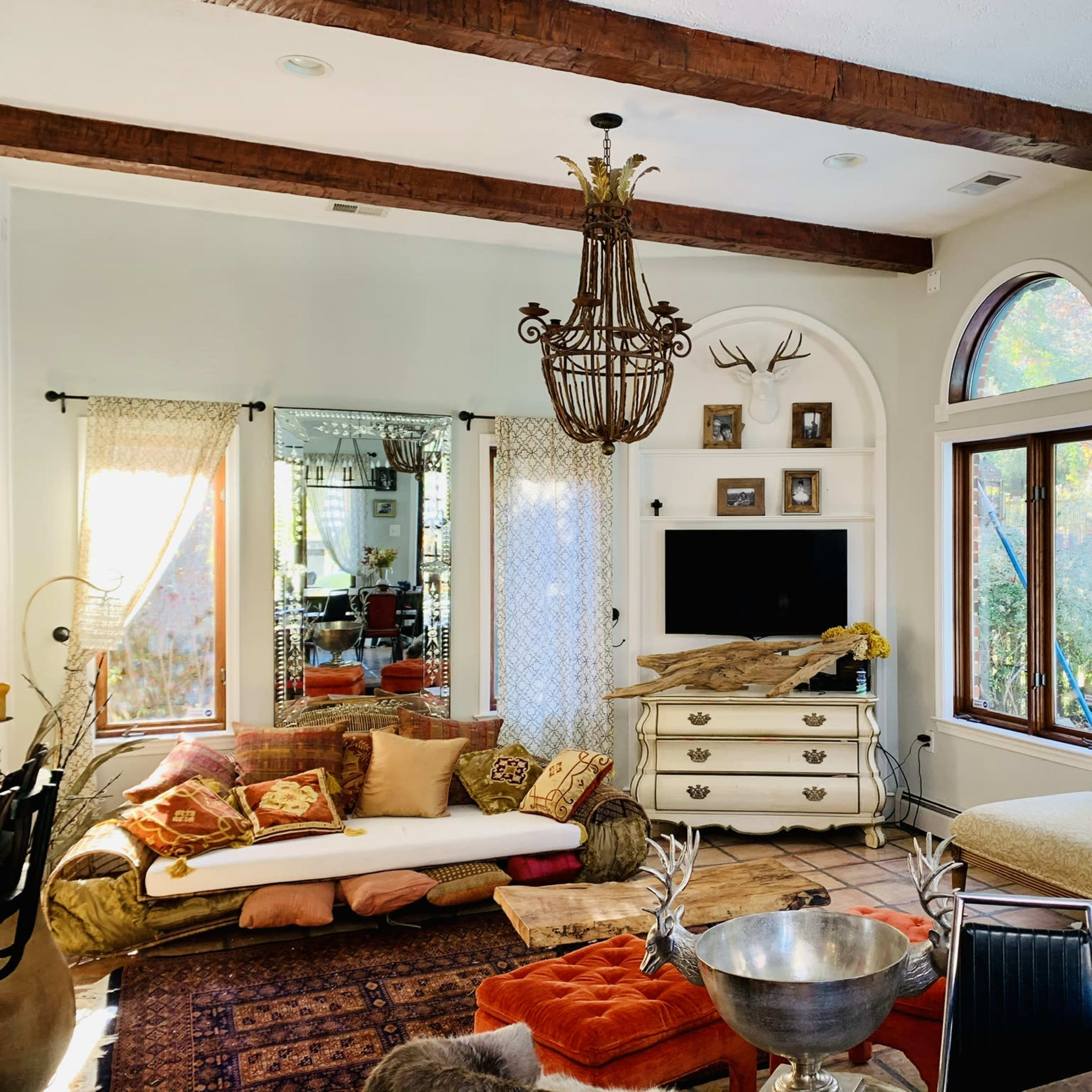 The spacious living room features large windows, a rustic chandelier, a comfortable seating area with decorative pillows, and a central entertainment unit.