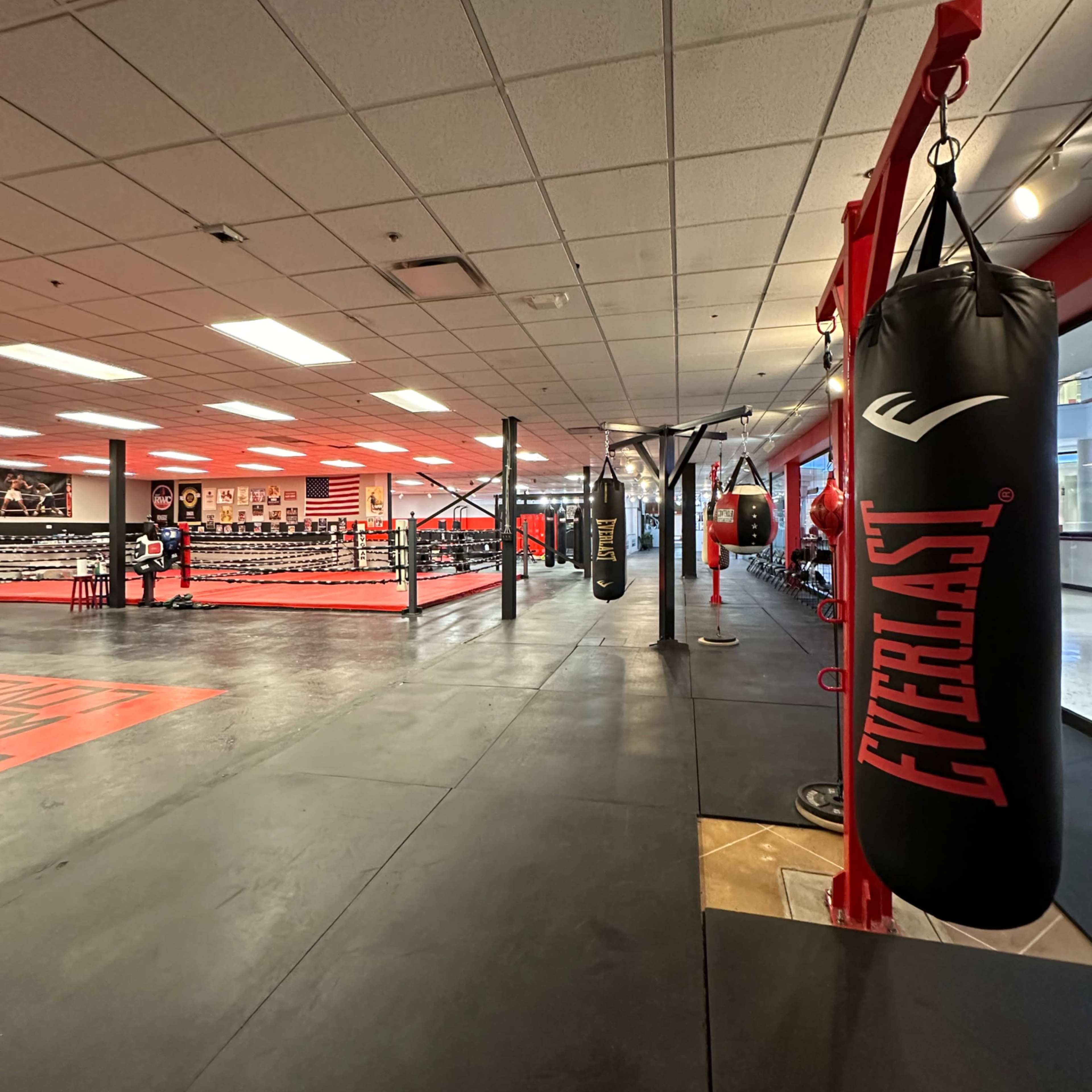 An interior view of a boxing gym featuring a boxing ring, several heavy bags, and a training area with rubber flooring.
