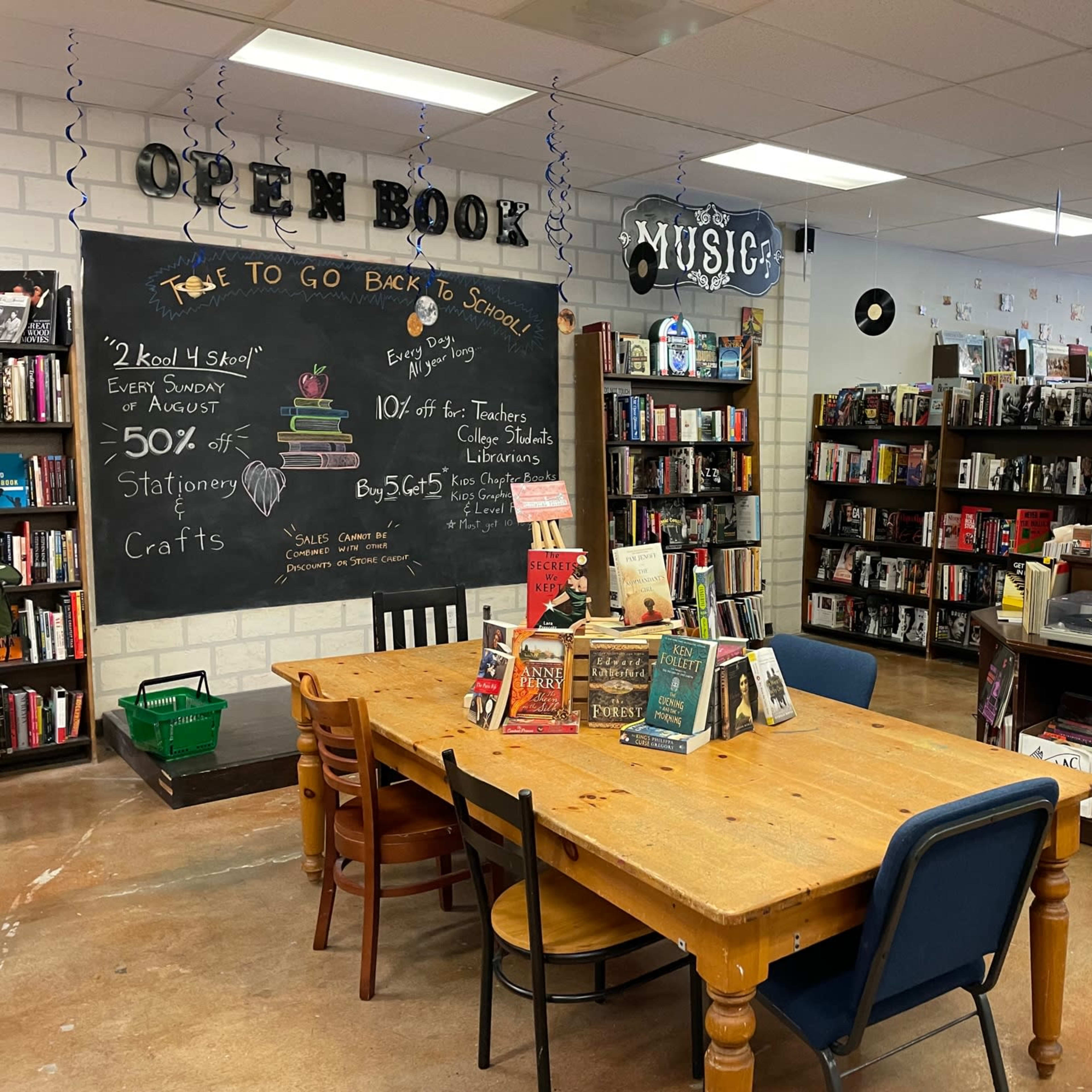 A cozy bookstore features wooden tables, bookshelves filled with various titles, and a chalkboard advertising promotions.