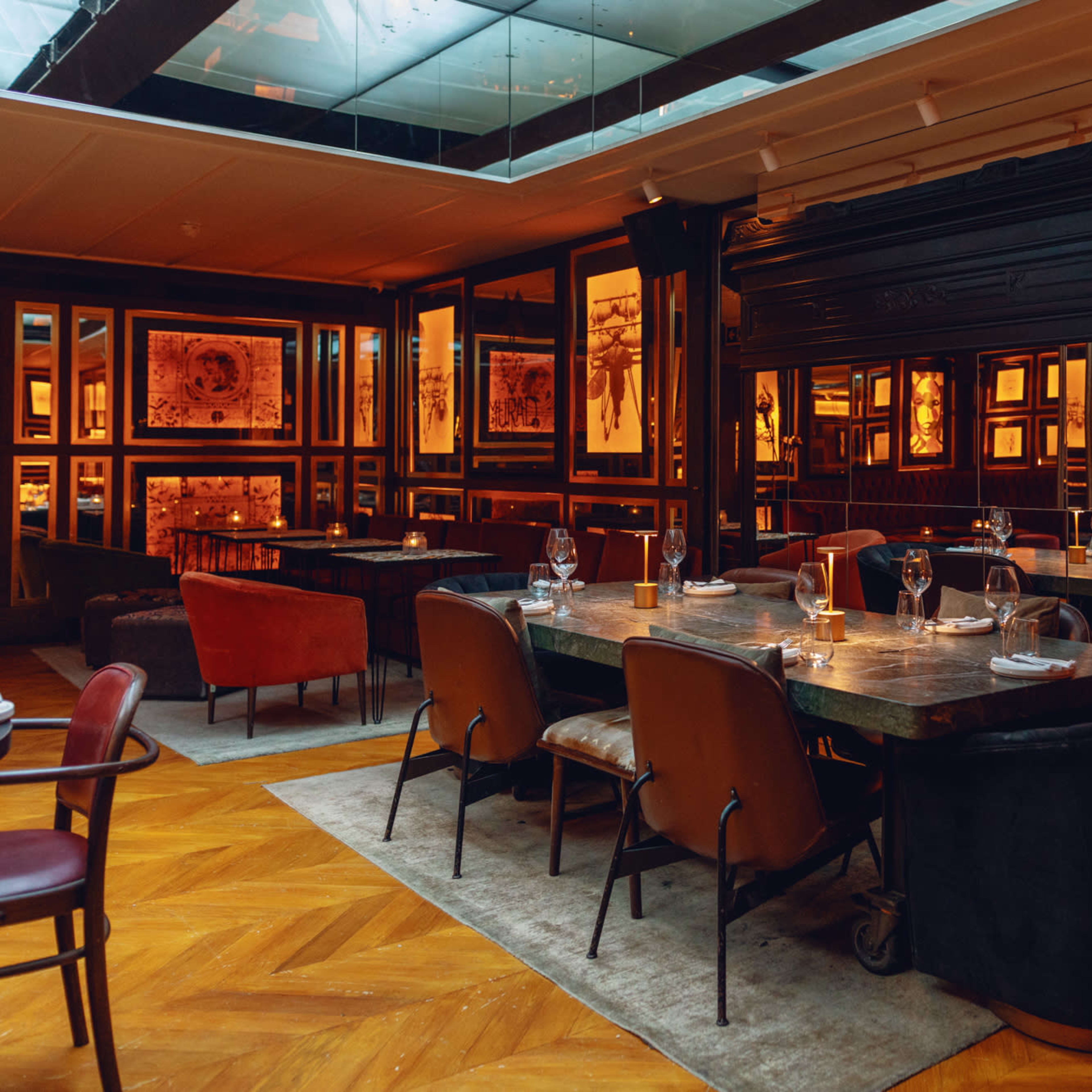 A warmly lit restaurant interior with wooden flooring, framed artwork on the walls, and a mix of seating arrangements including chairs and tables.