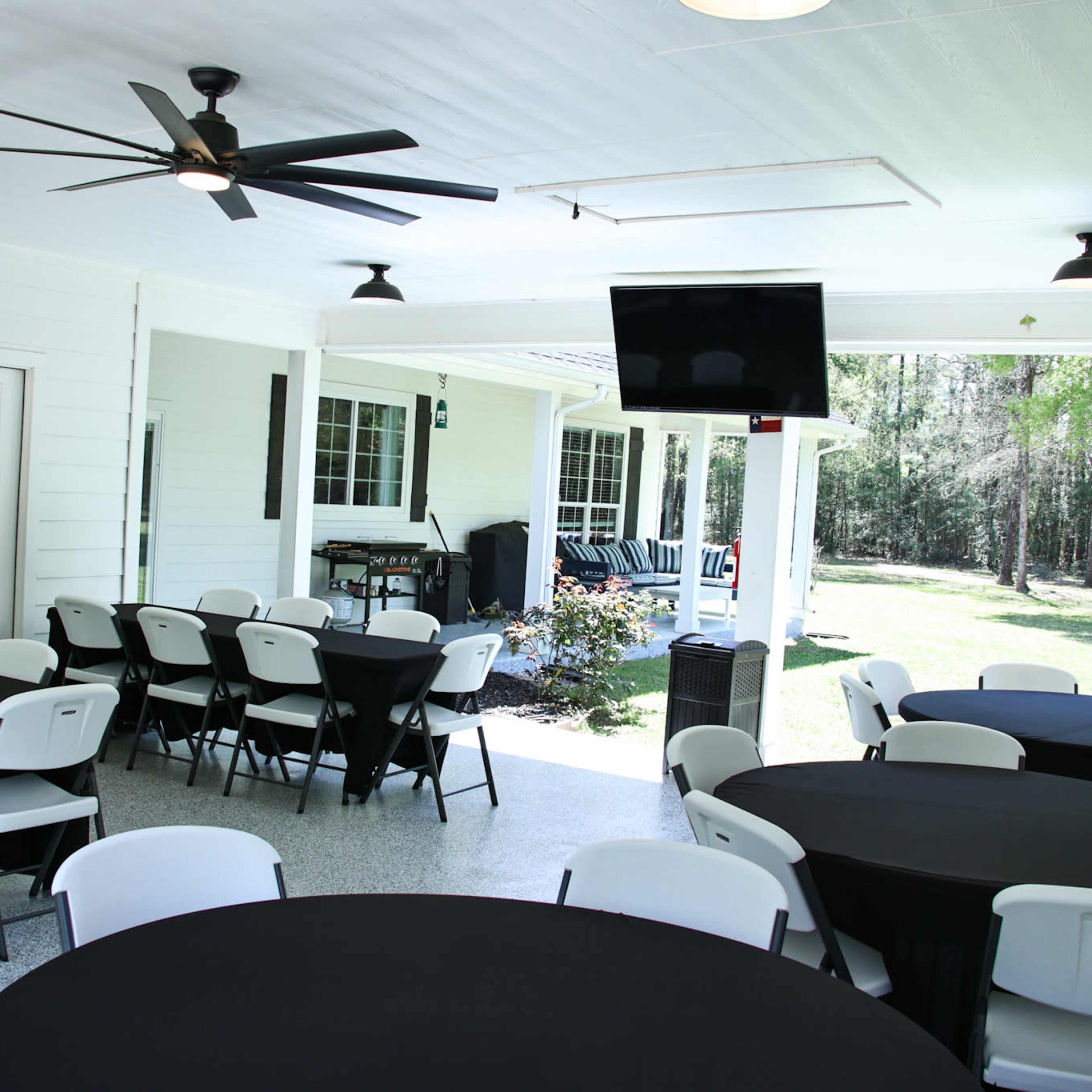 A spacious outdoor room with several round tables covered in black cloth, a ceiling fan, and a television mounted on the wall.