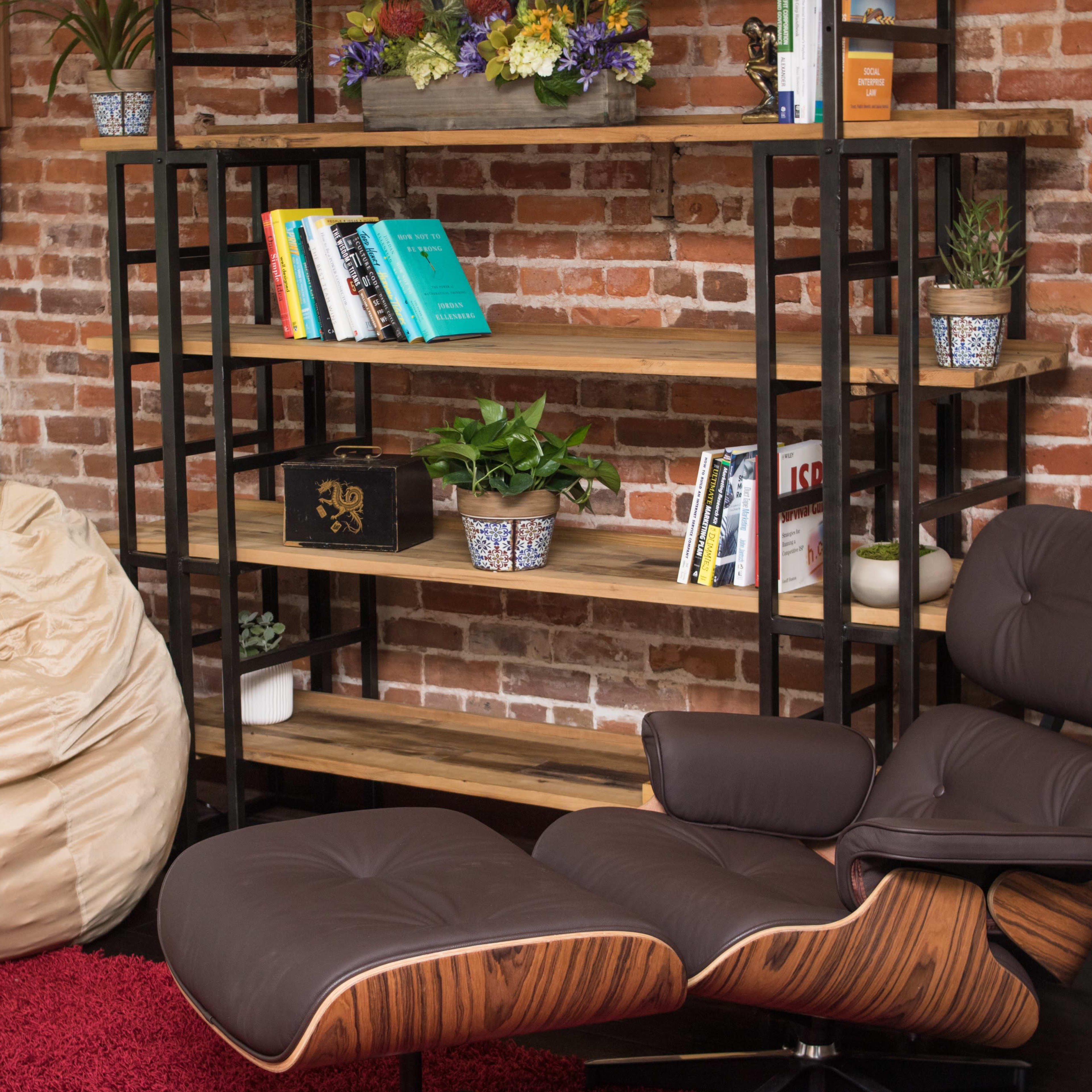 A cozy room featuring a brown leather chair next to a bookshelf filled with books and plants, with a tan bean bag on one side and a red rug on the floor.