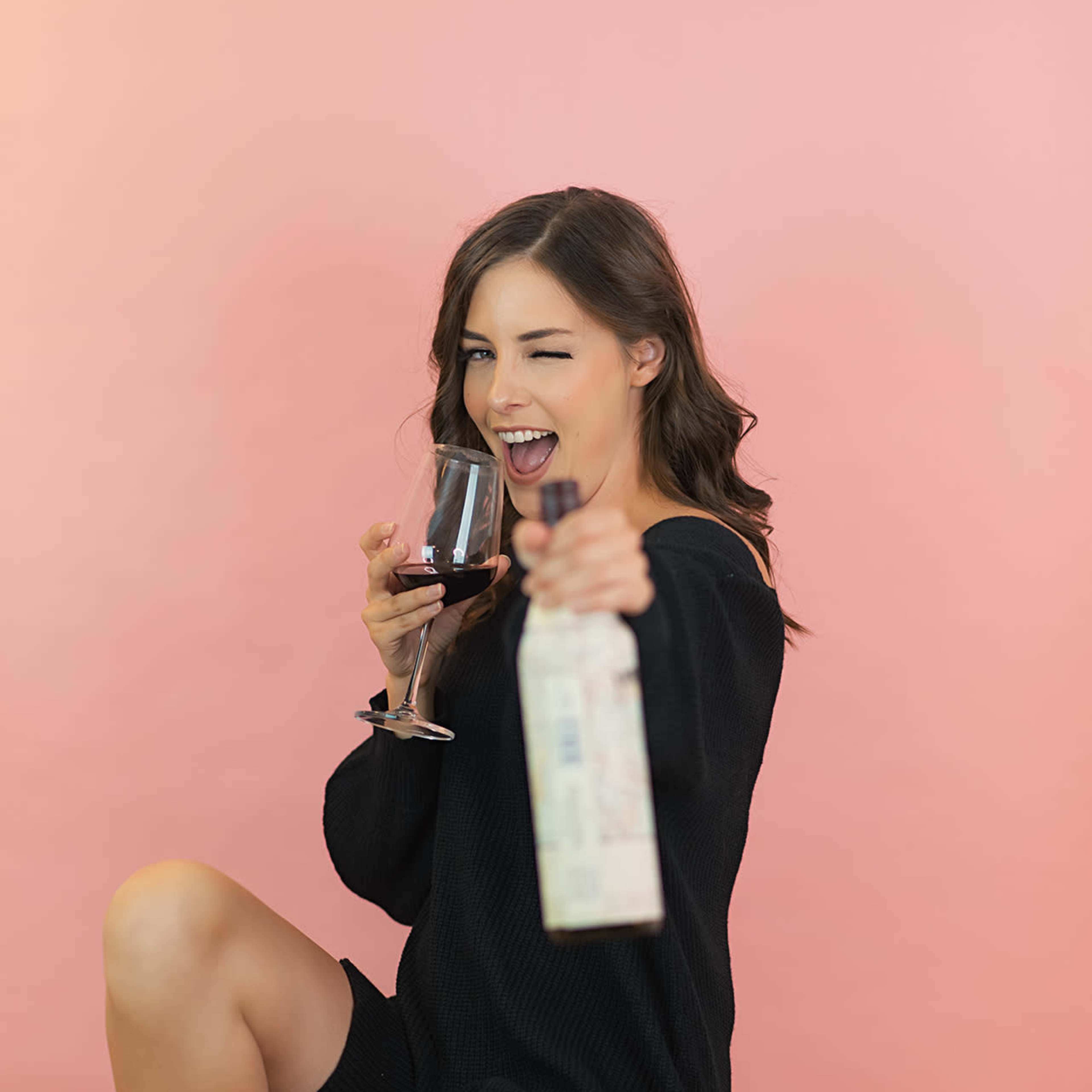 A woman in a black sweater holds a glass of red wine and a wine bottle while sitting on a stool against a pink background.