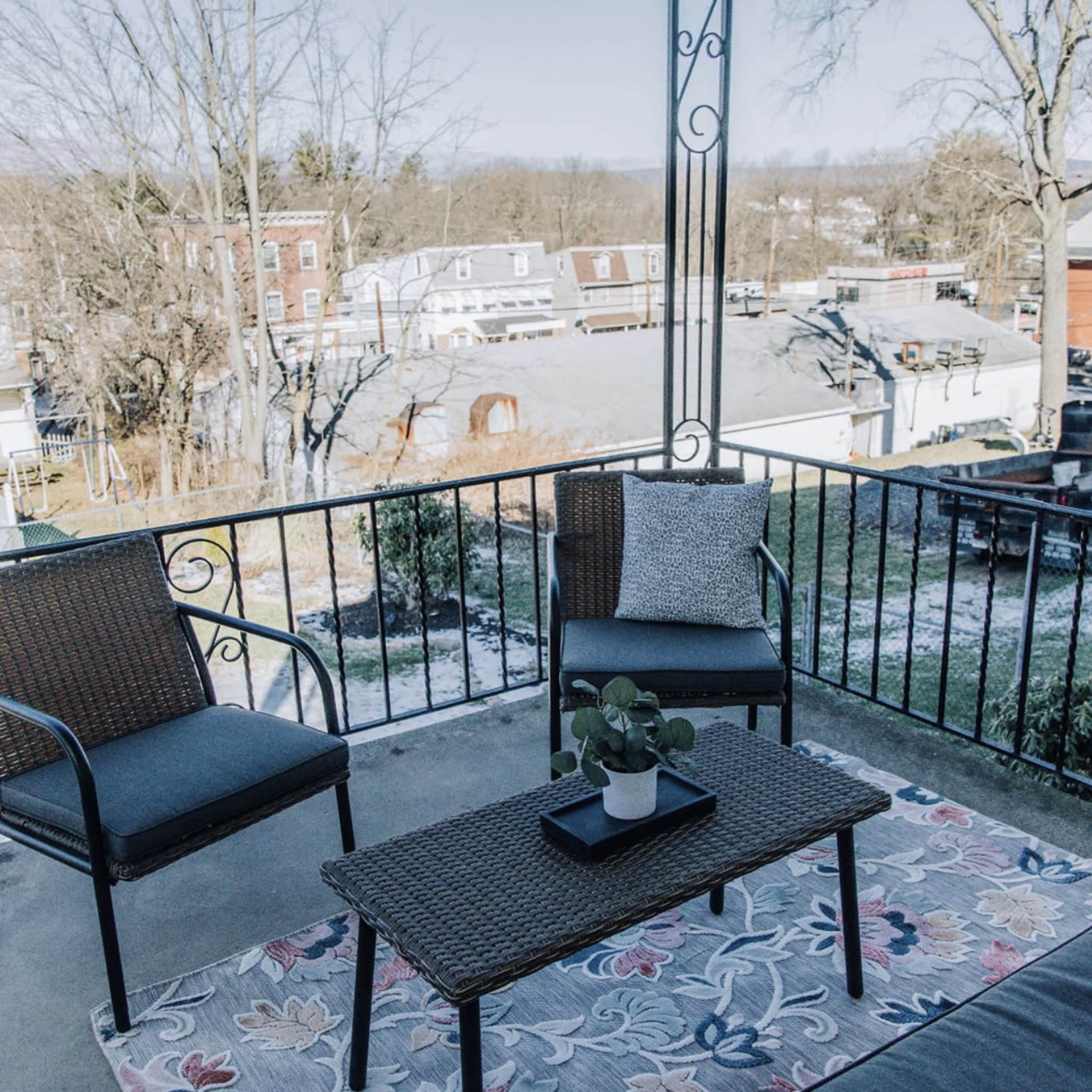 A small outdoor patio area with two chairs and a table on a patterned rug, overlooking a residential neighborhood.