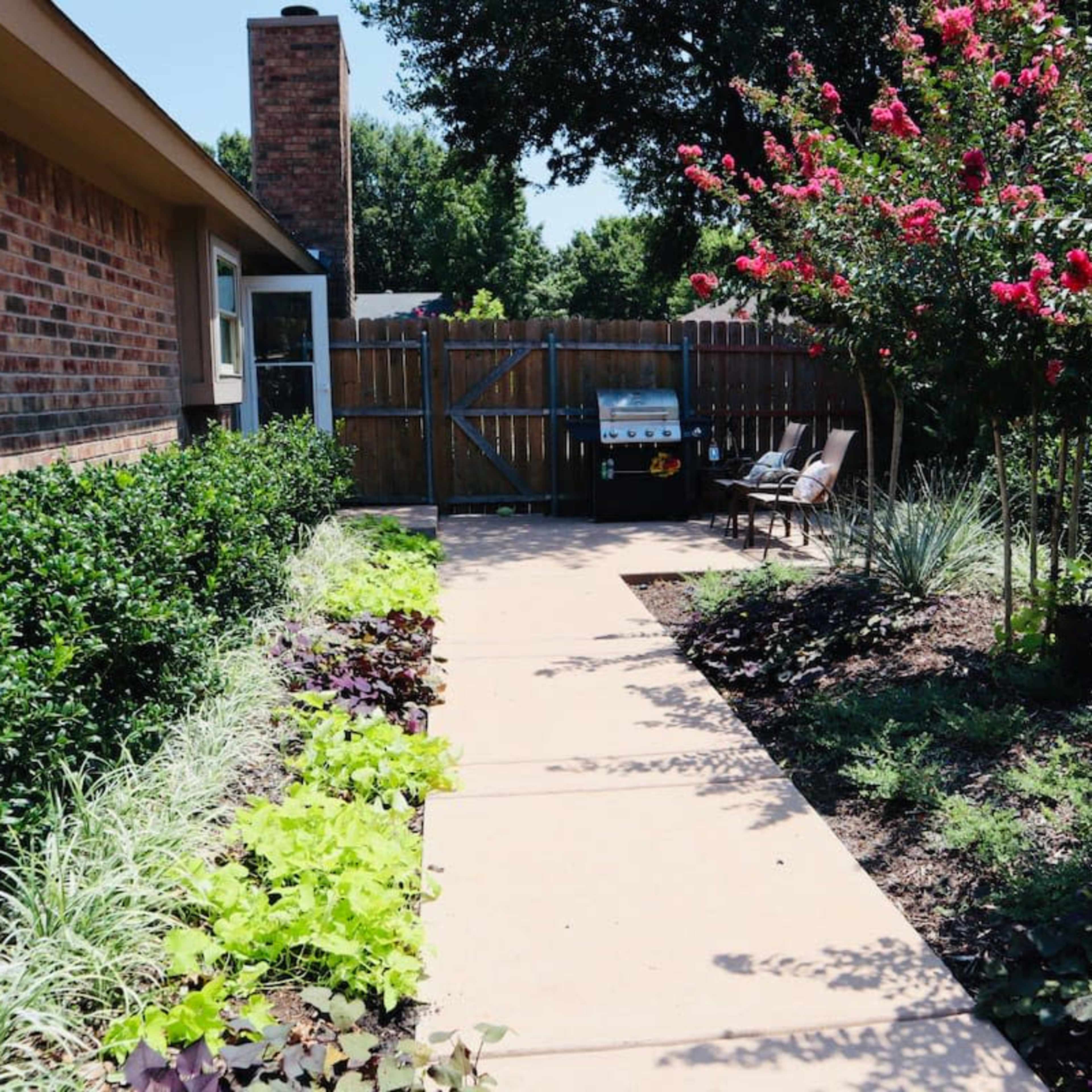 A concrete walkway leads through landscaped gardens to a fenced area with a grill and seating.