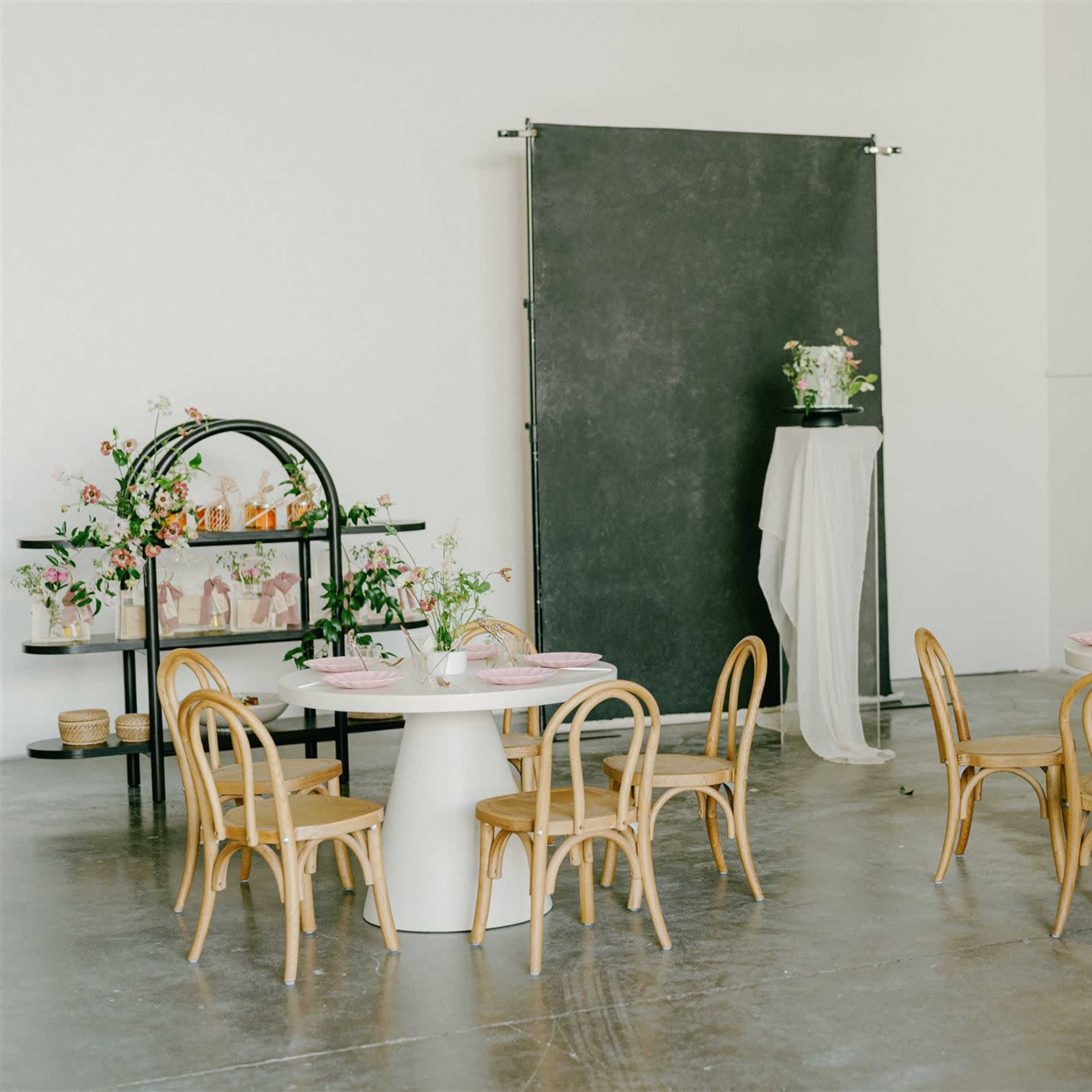 A spacious, well-lit room features two round tables with wooden chairs, a decorative shelf with flowers, and a backdrop made of dark fabric.