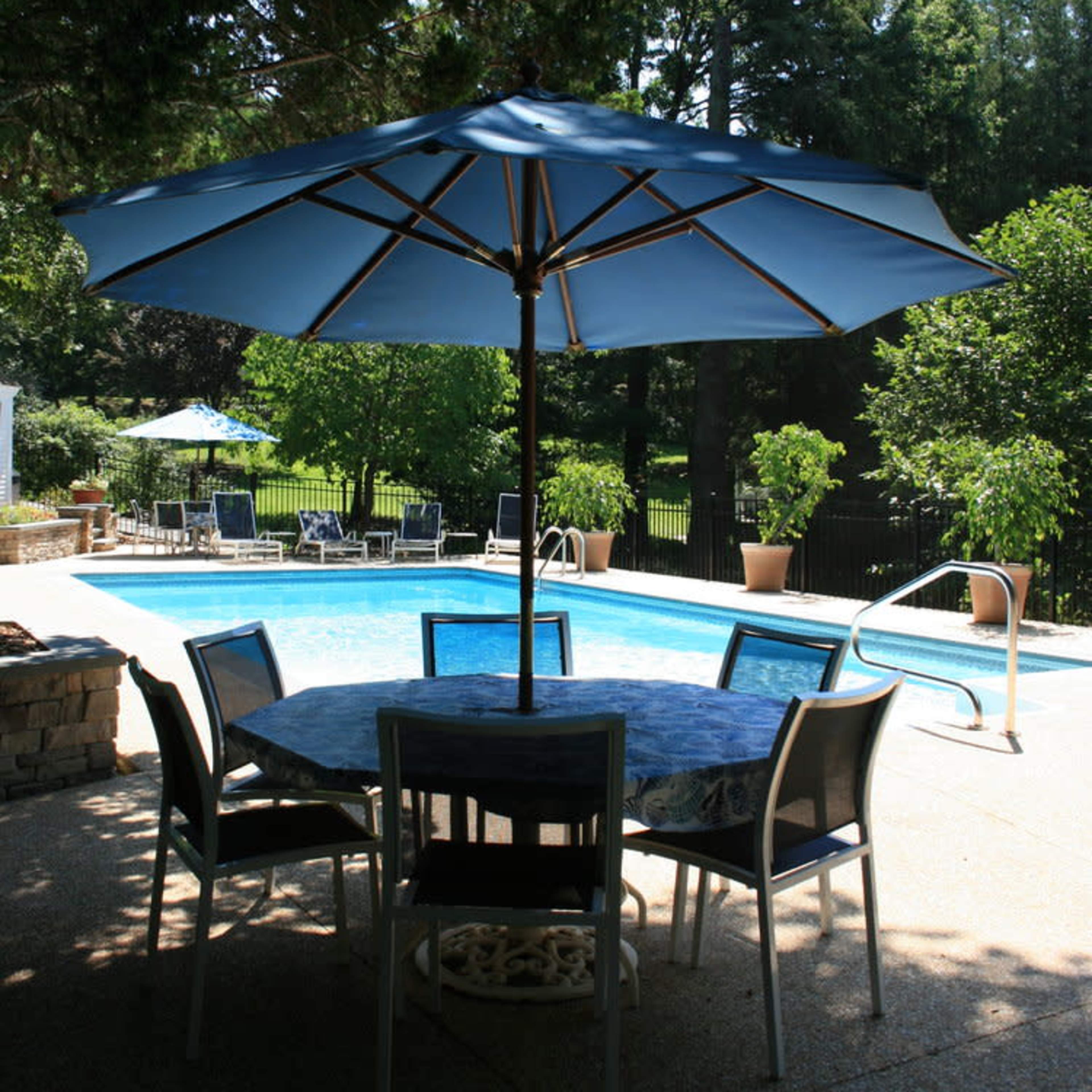 A blue umbrella shades a round table surrounded by chairs near a swimming pool in a backyard.