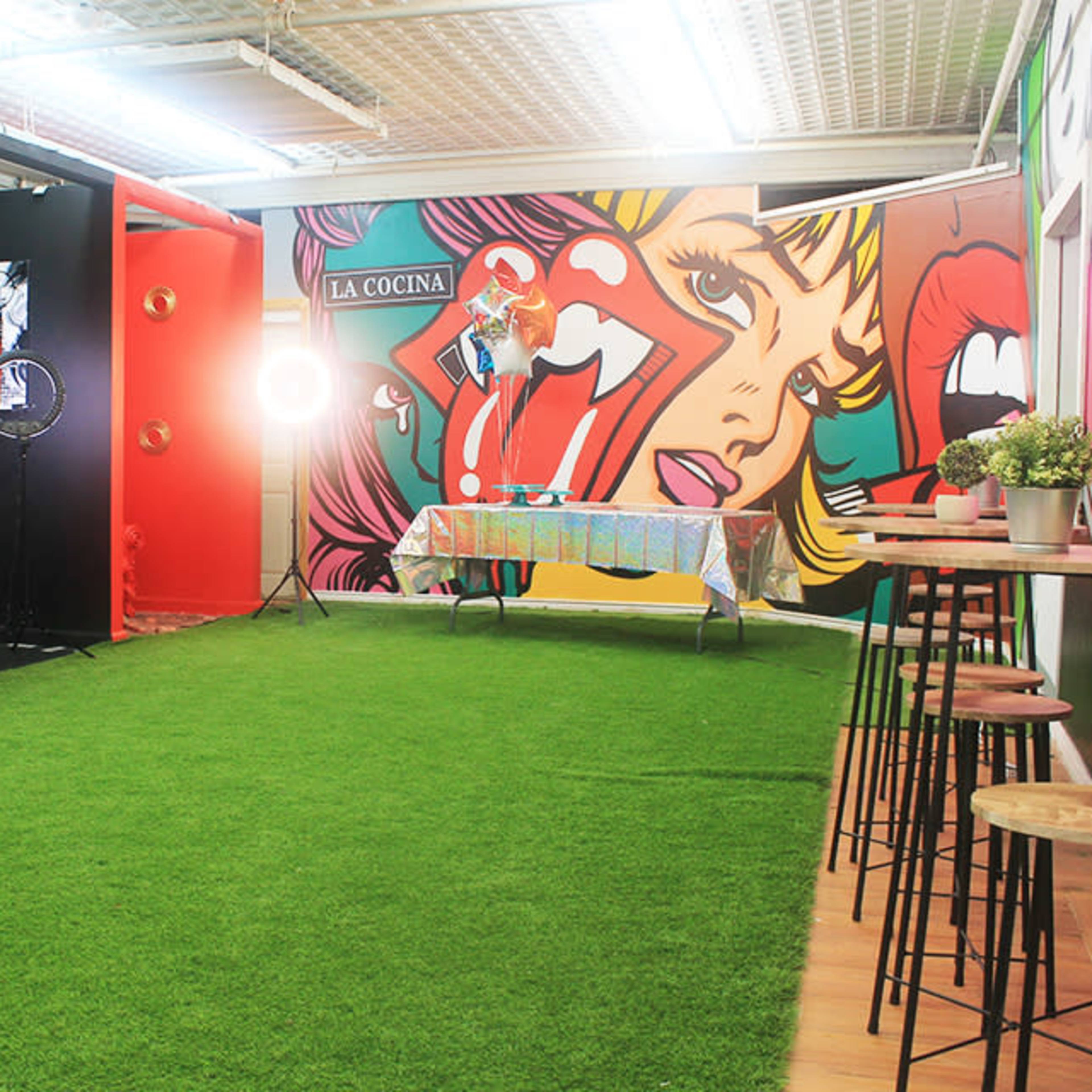 A brightly colored interior space with green artificial grass flooring, featuring a decorated table, a chair, and a vibrant mural on the wall.