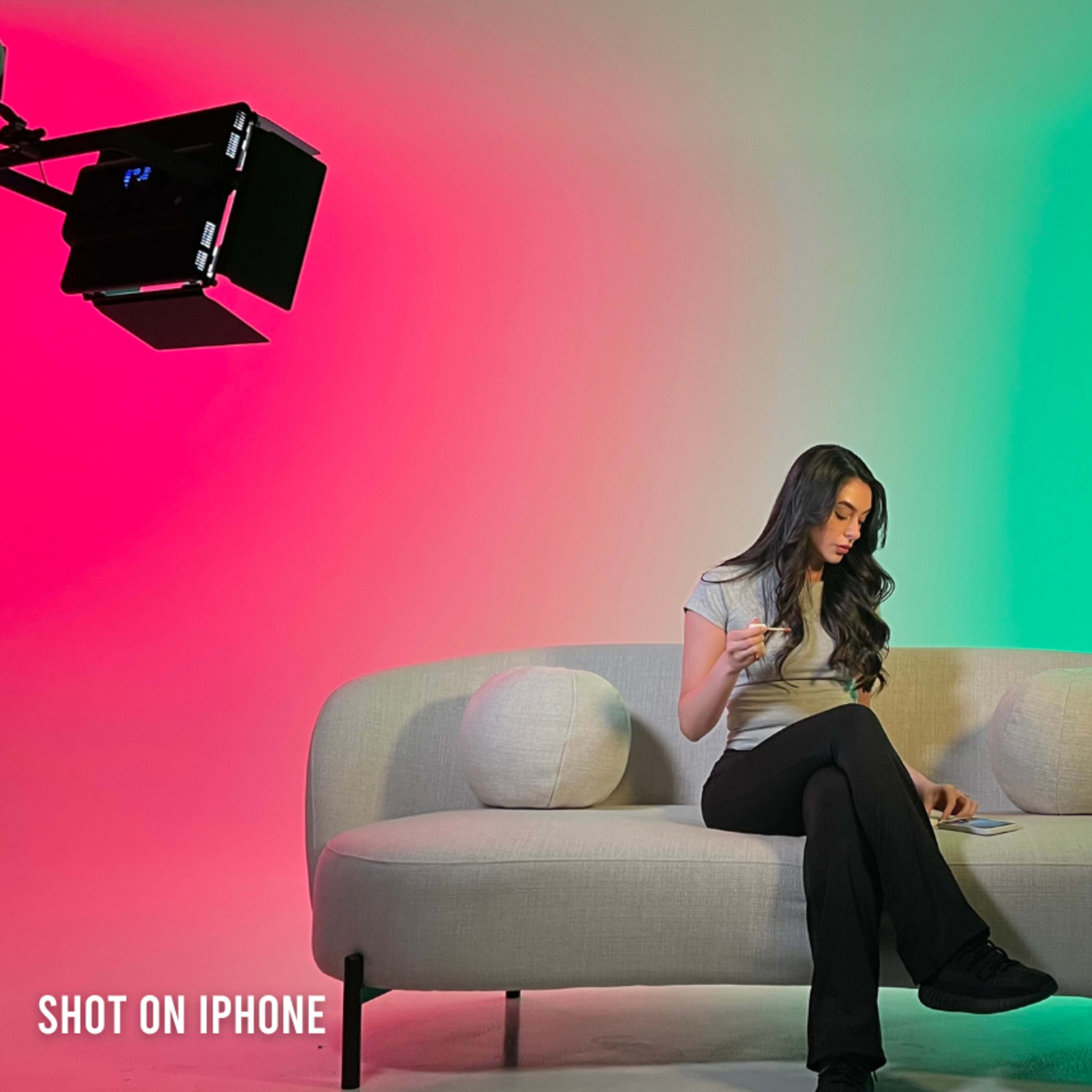 A woman sits on a light-colored sofa, holding an object, with colorful lighting in the background.
