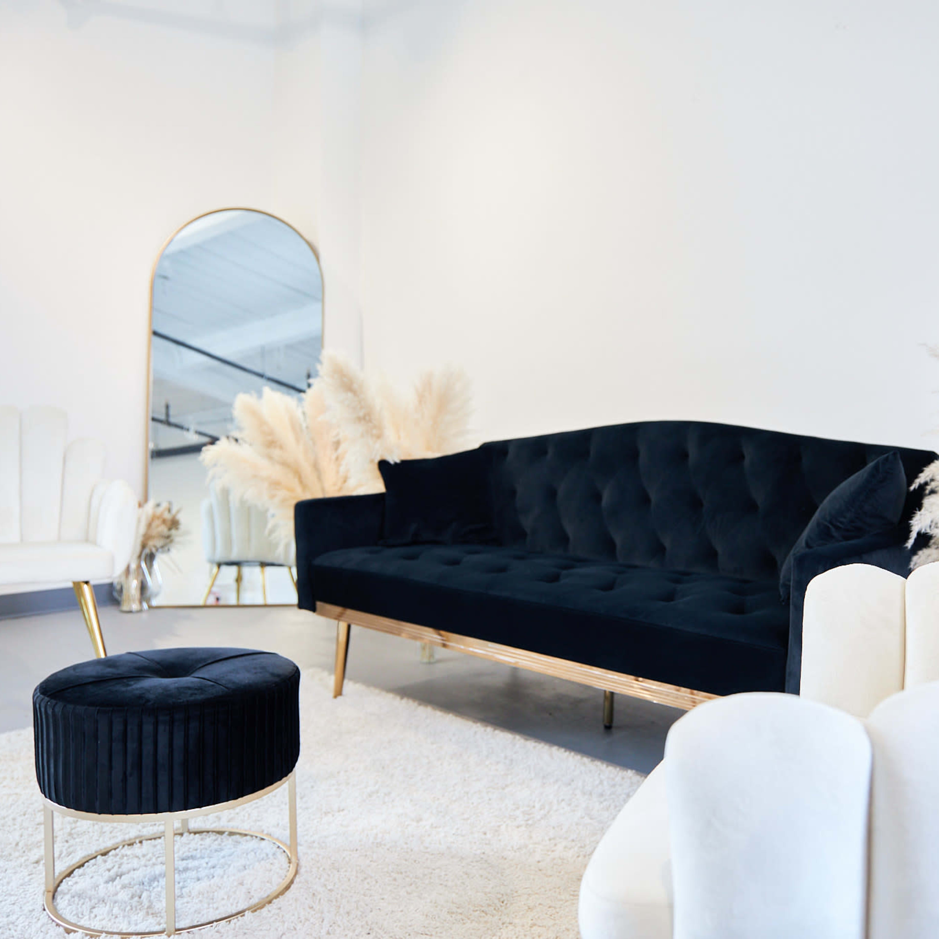 A modern interior features a black velvet sofa, white upholstered armchairs, a round ottoman, and decorative pampas grass arranged in the background.