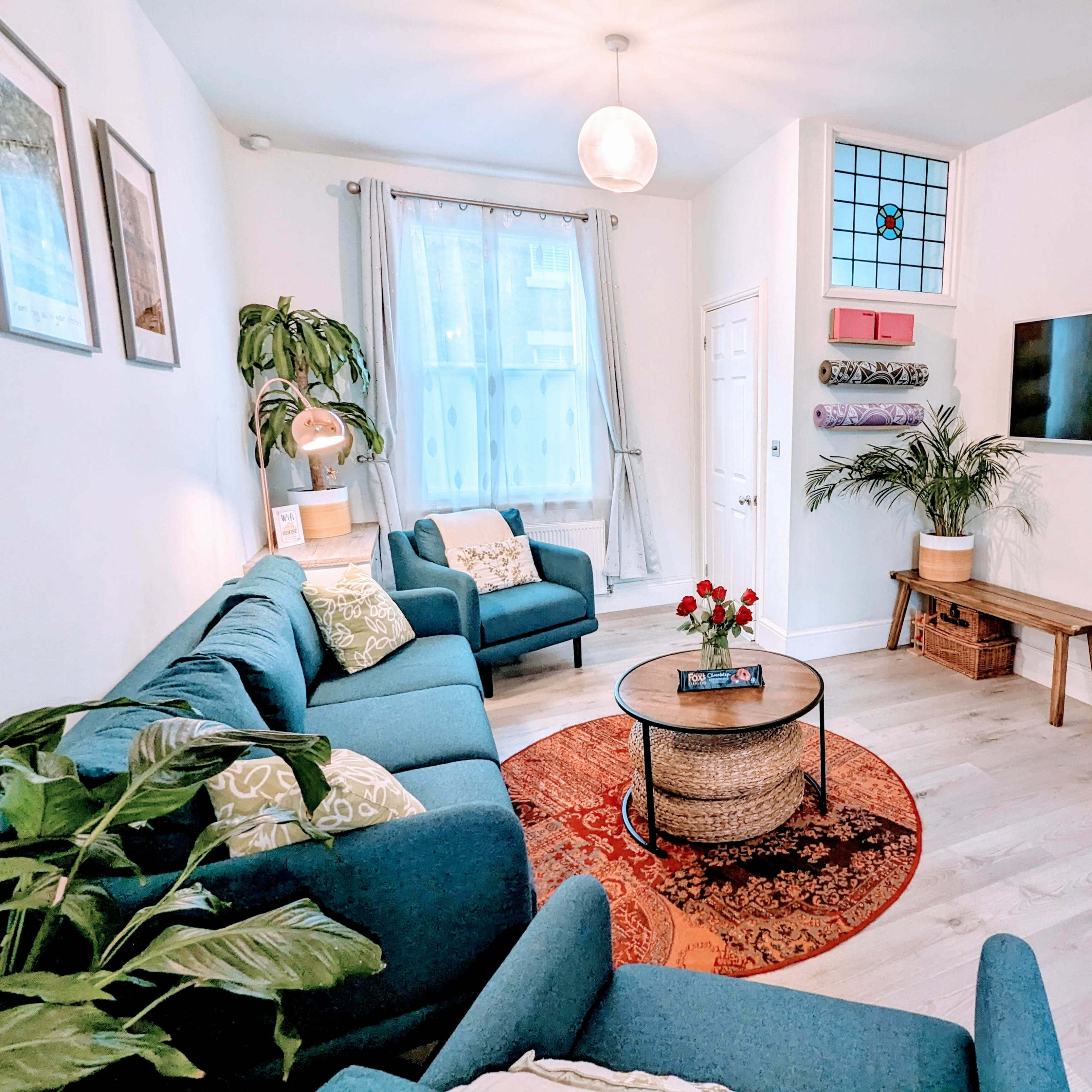 The living room features a blue sectional sofa, a round coffee table with a floral centerpiece, and a wall-mounted TV against a bright, airy background.