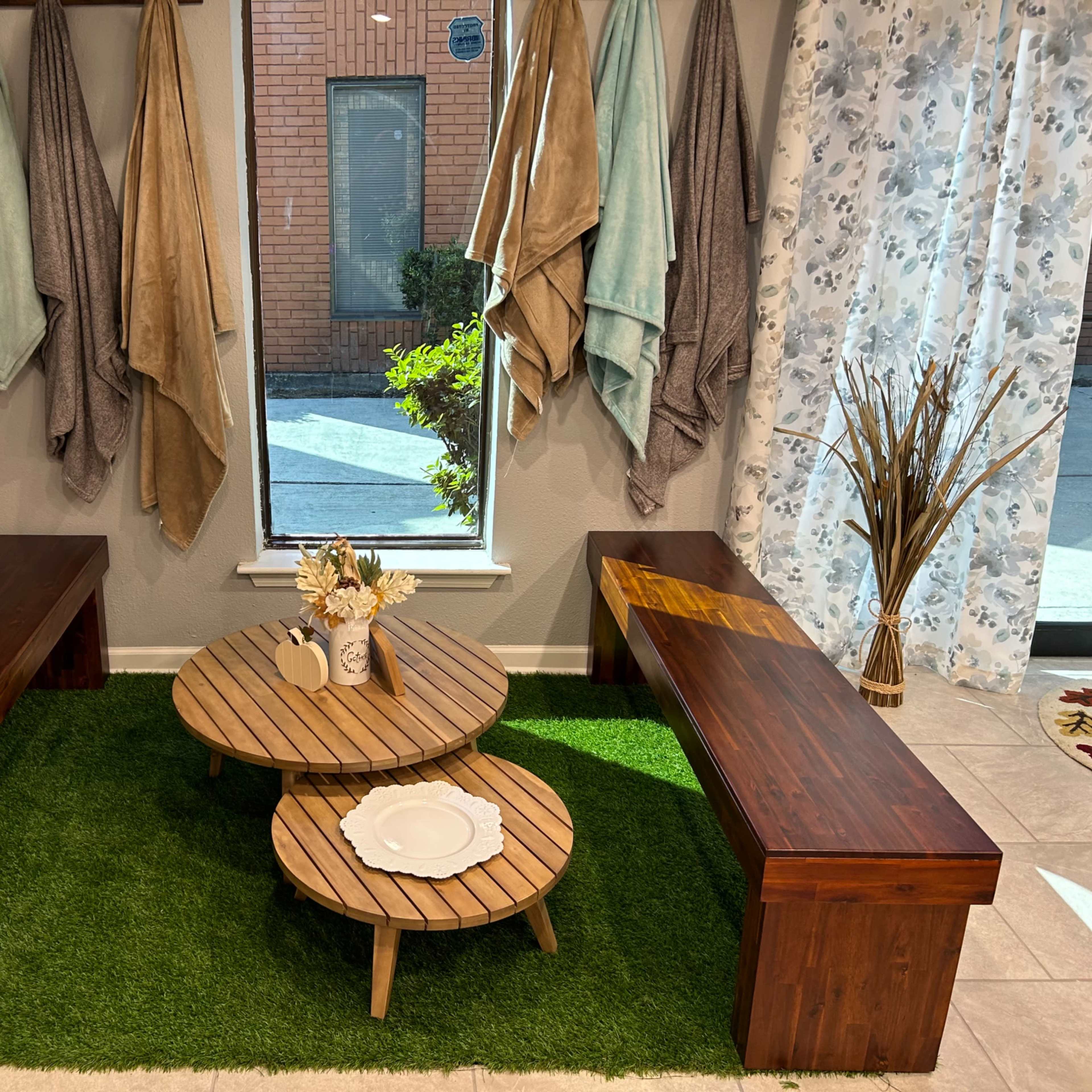A cozy corner with two wooden benches, a circular wooden coffee table, and decorative plants on green carpet flooring near a large window.
