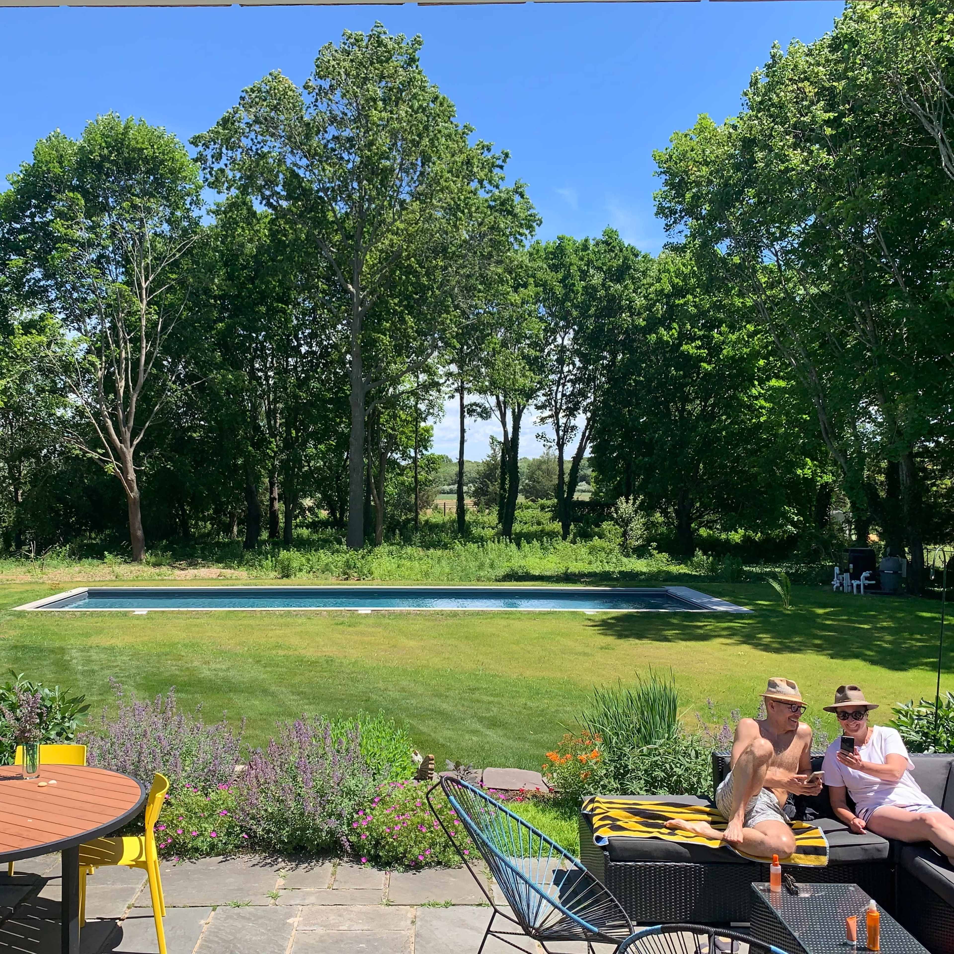 A group of people relaxes near a swimming pool surrounded by trees and a landscaped garden.