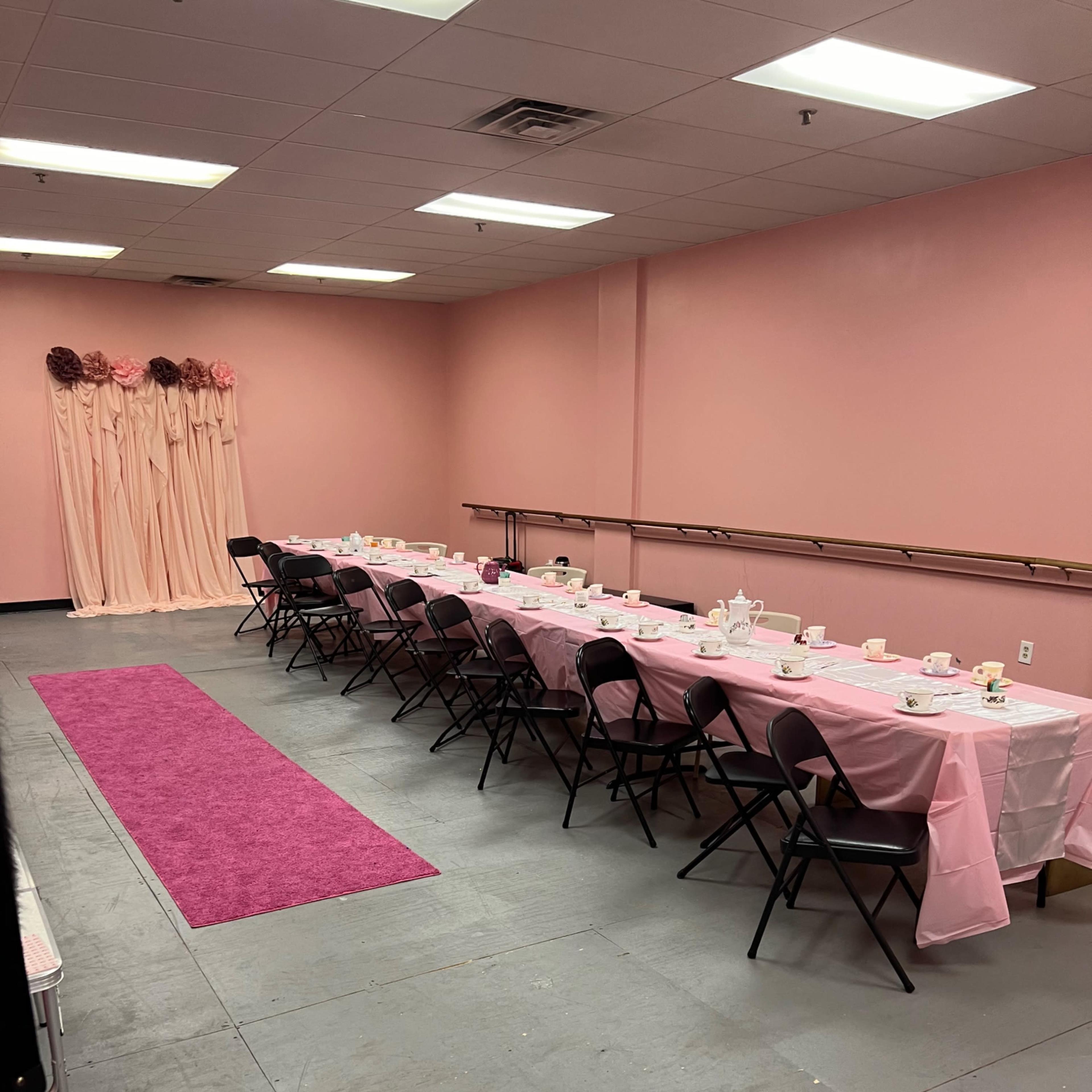 A pink-themed banquet room set with a long table adorned with a pink tablecloth, black chairs, and tea service items, accompanied by a pink carpet and decorative backdrop.