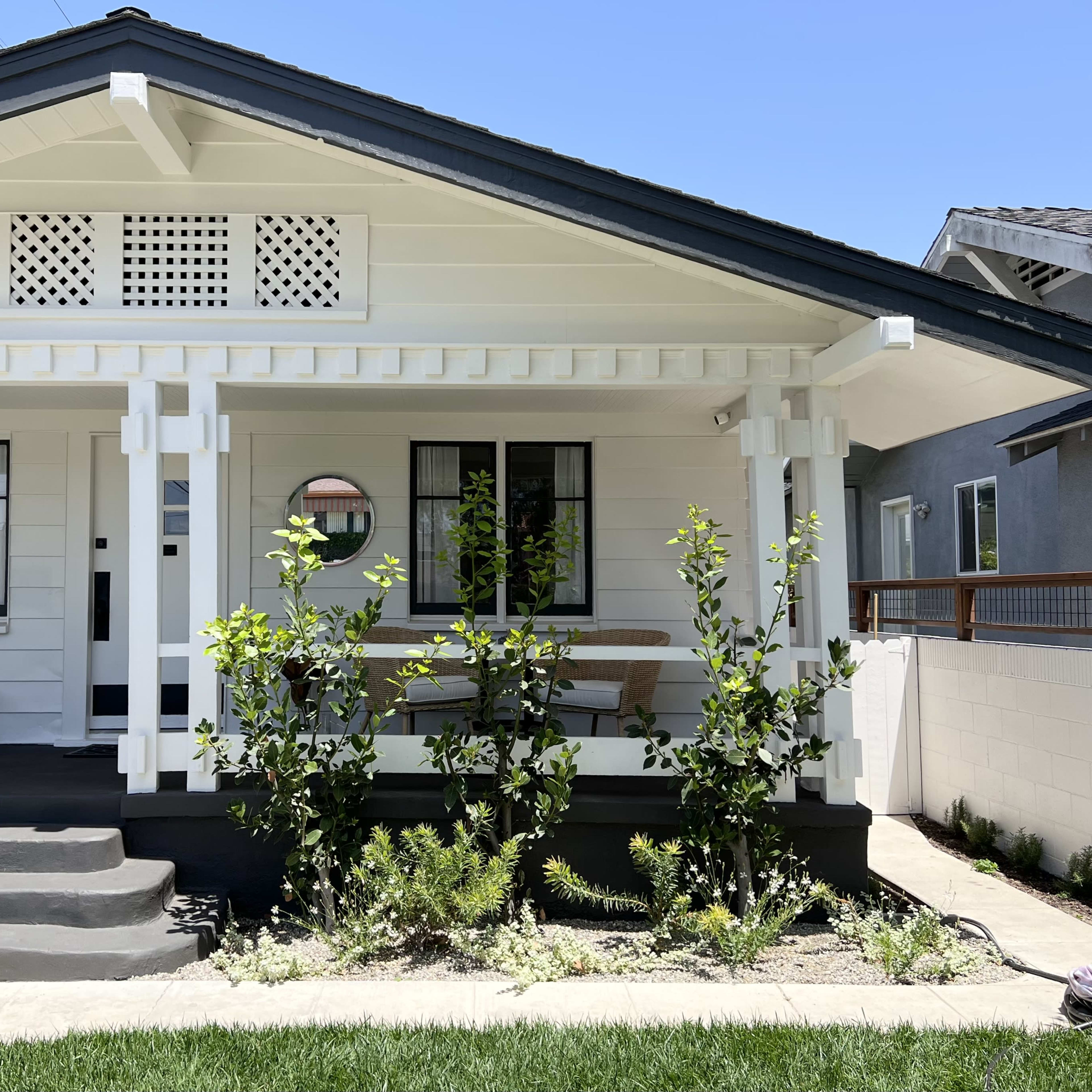 A white house with a porch, black accents, and greenery in the front yard under a clear blue sky.