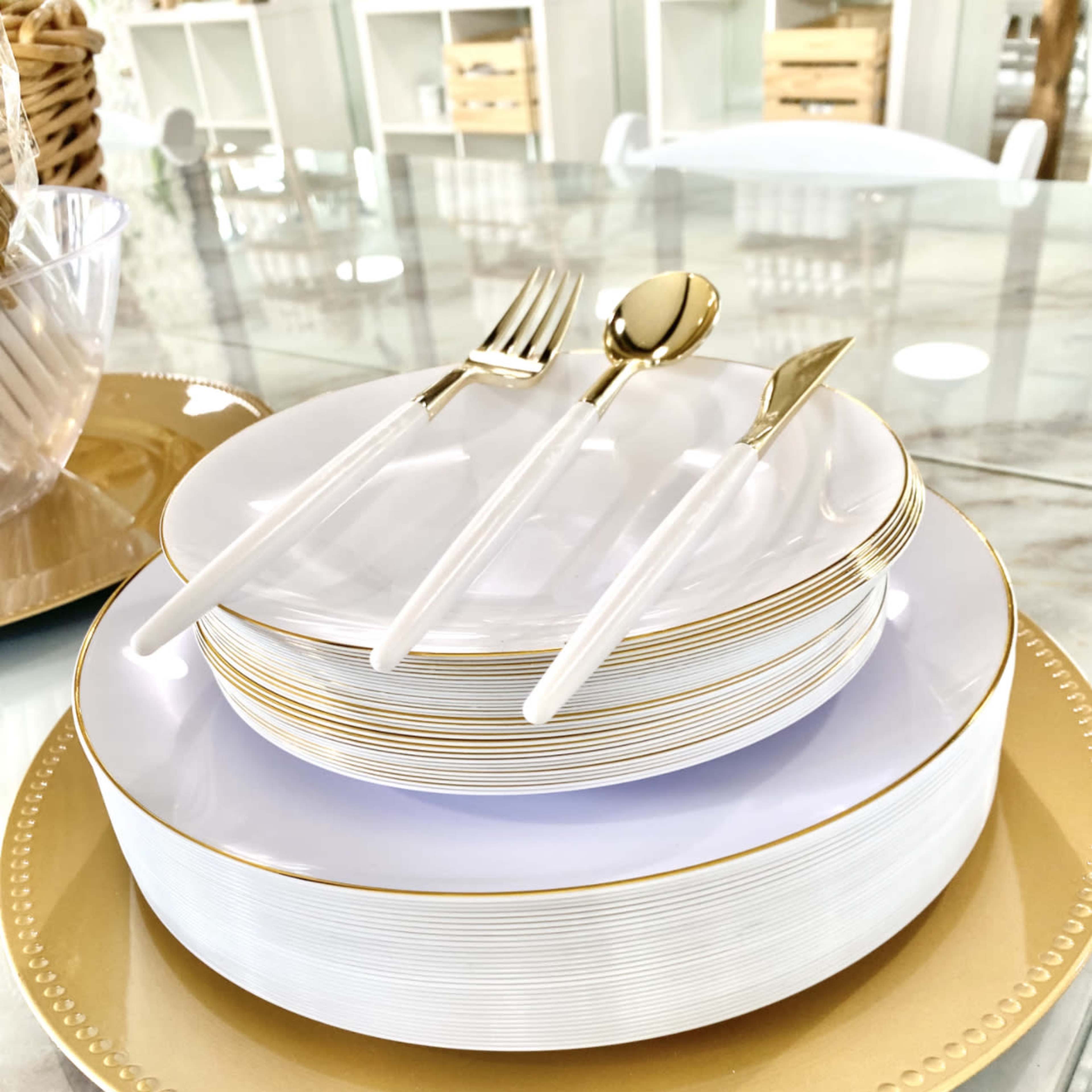 A set of stacked white plates with gold trim and silverware is arranged on a gold charger plate on a glass table.