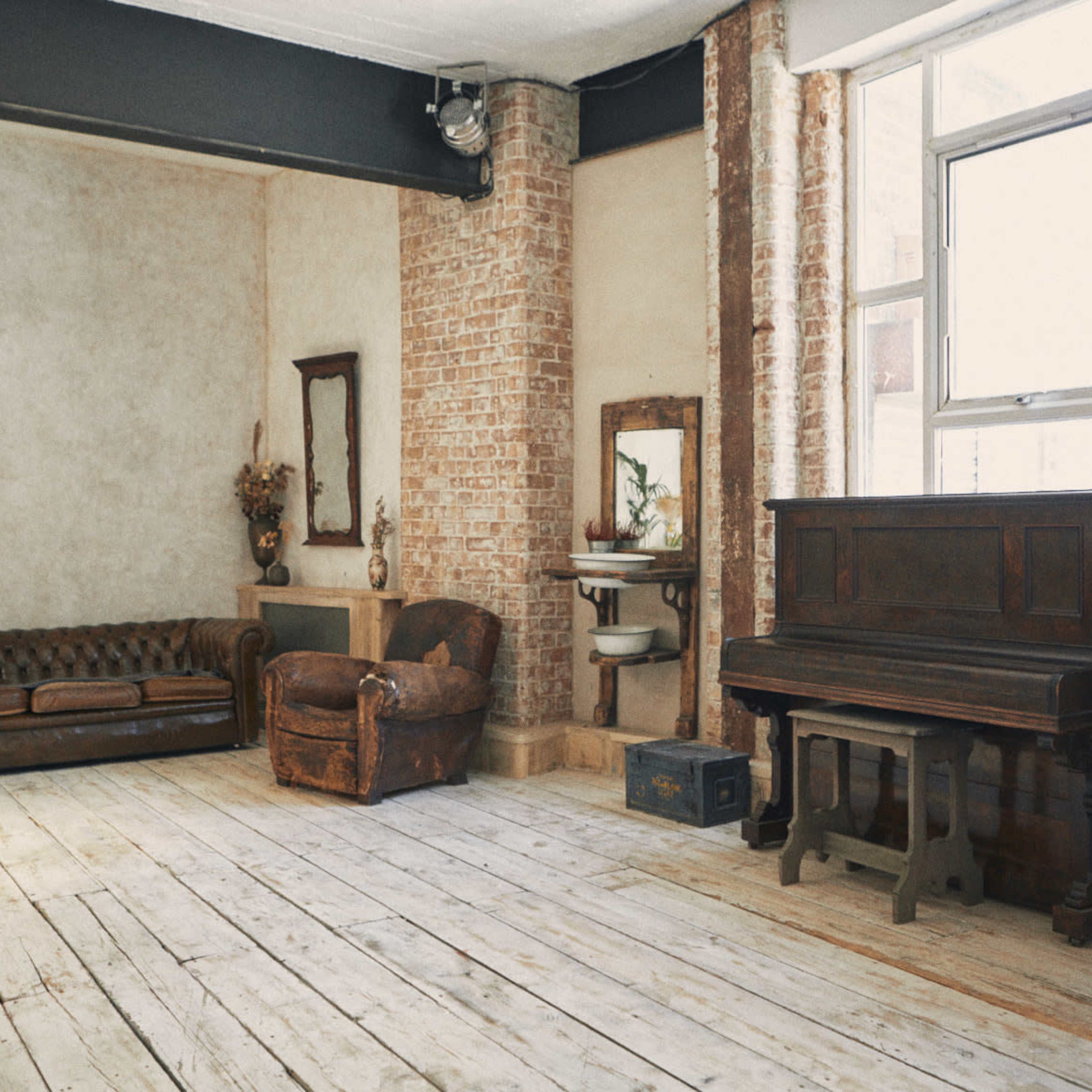 The room features a wooden floor, exposed brick walls, a vintage leather sofa, an antique armchair, and a piano.