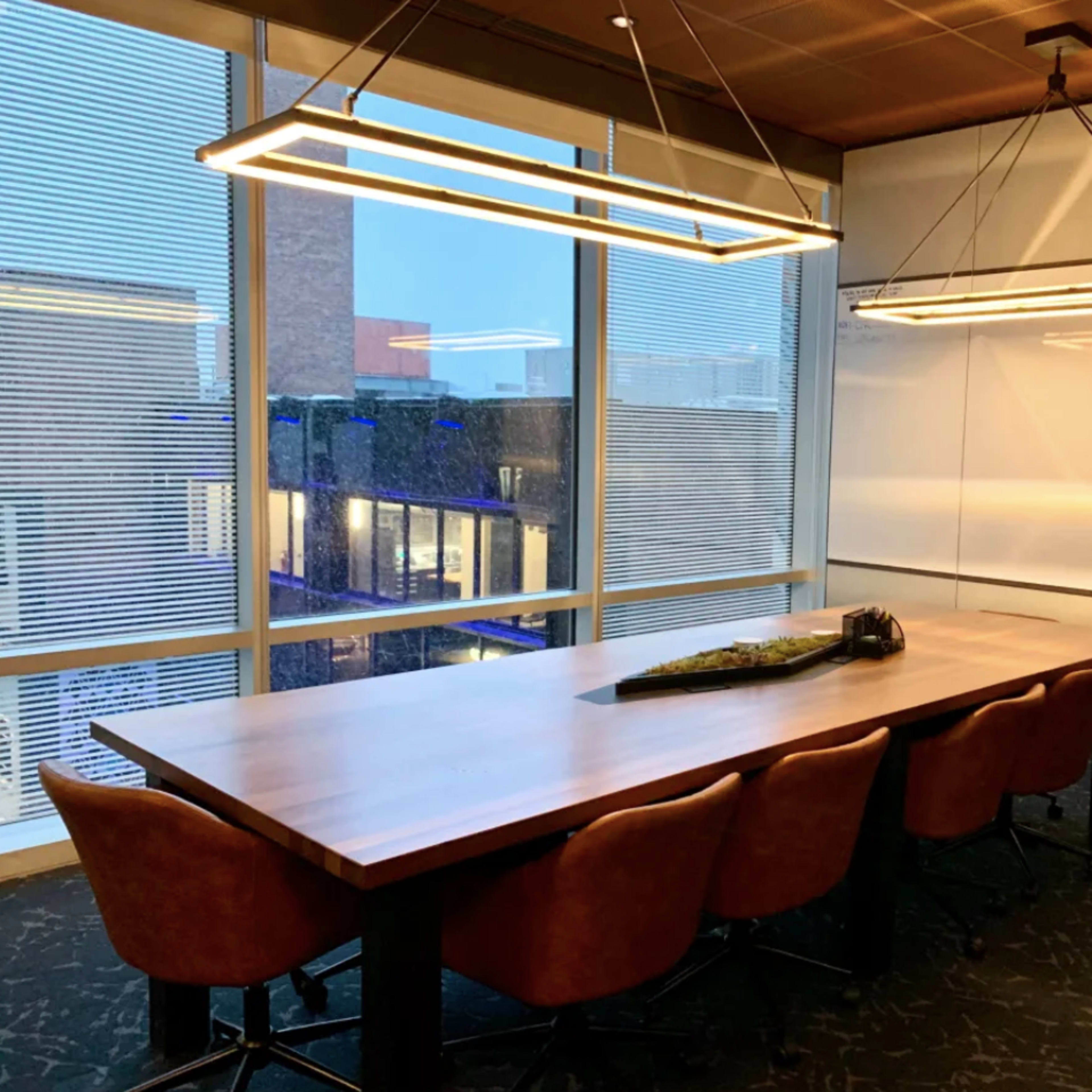A modern conference room features a large table surrounded by brown upholstered chairs and illuminated by two linear pendant lights.