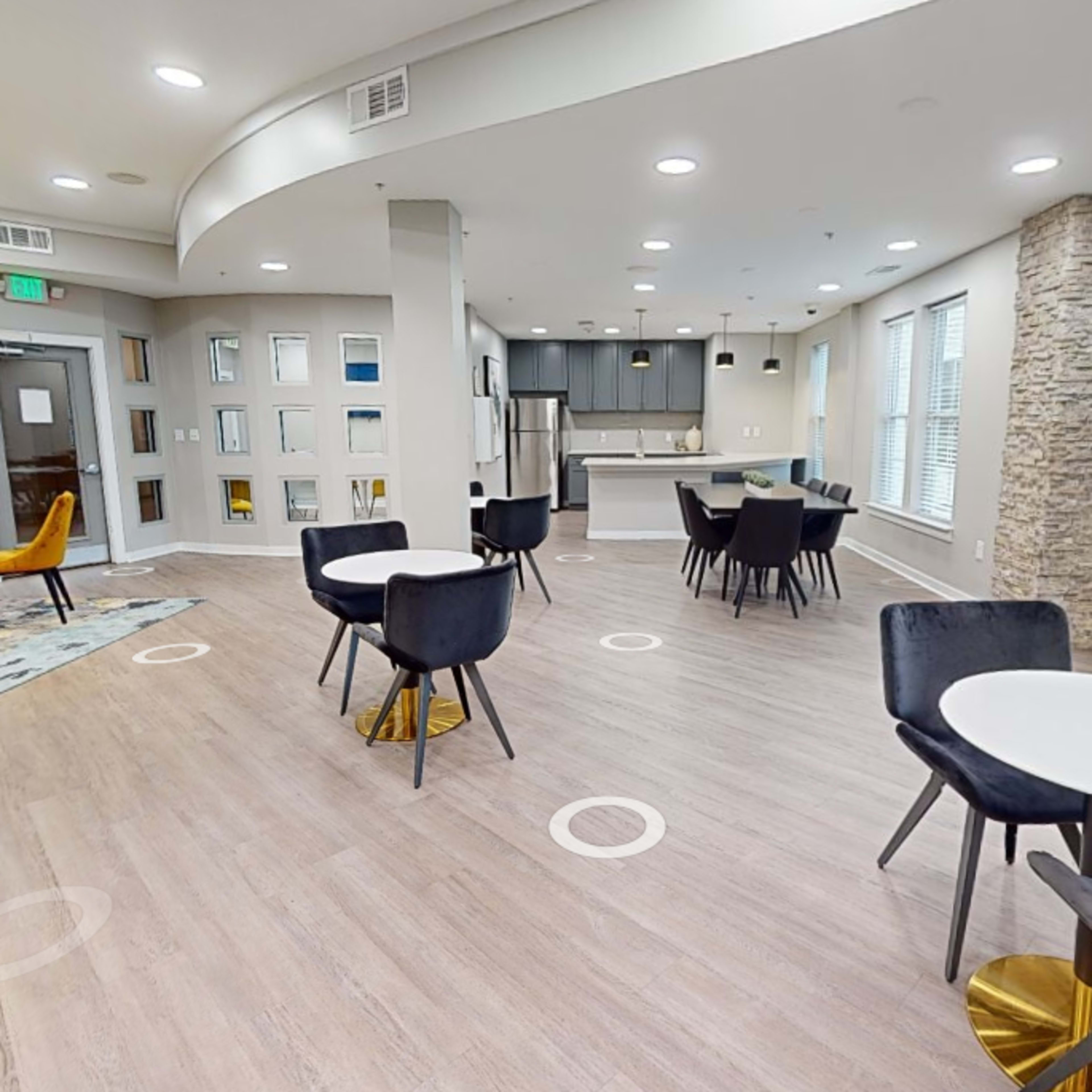 A modern lounge area with seating arrangements, including tables and chairs, and a kitchenette in the background.