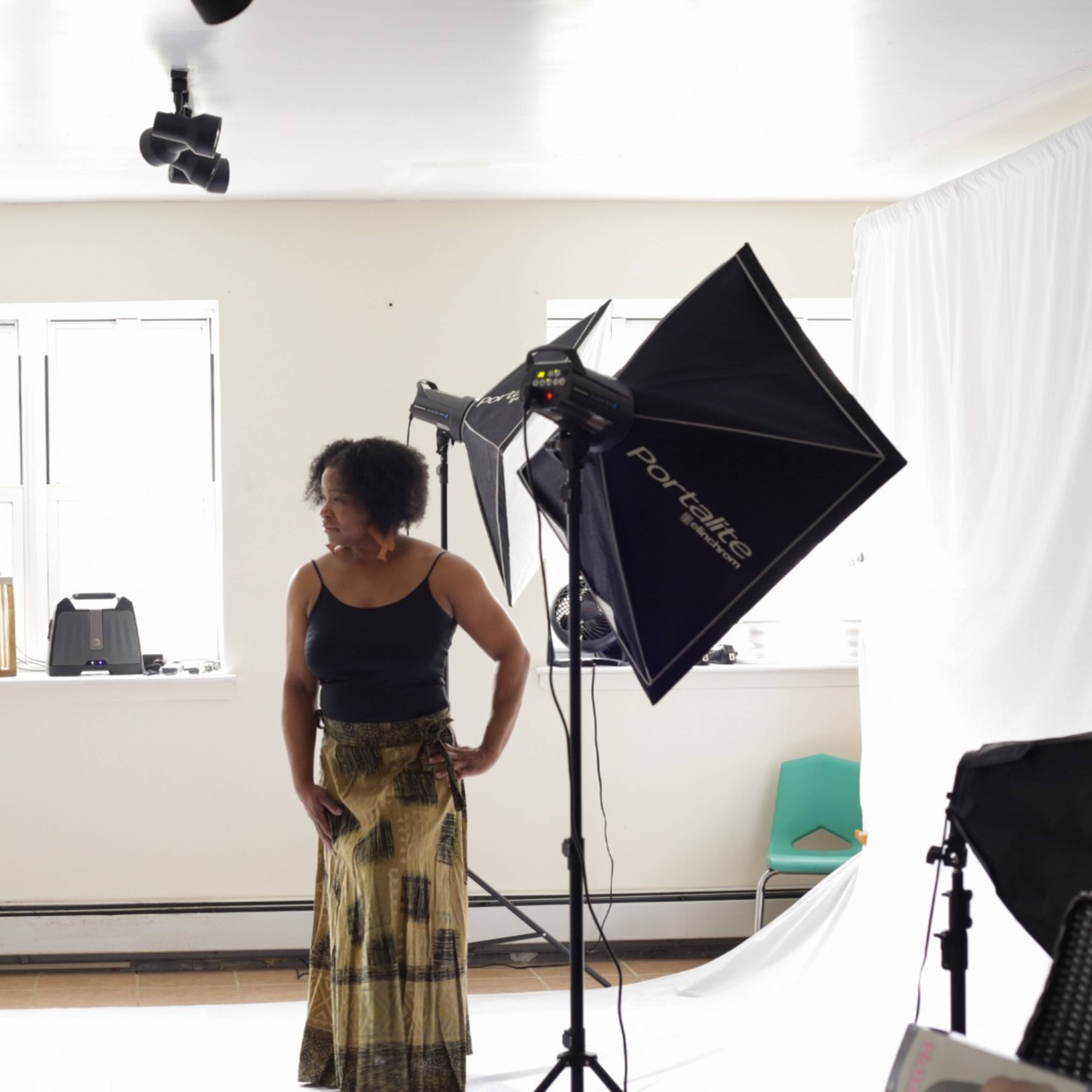 A woman stands posing in a brightly lit photography studio while a man adjusts his camera equipment.