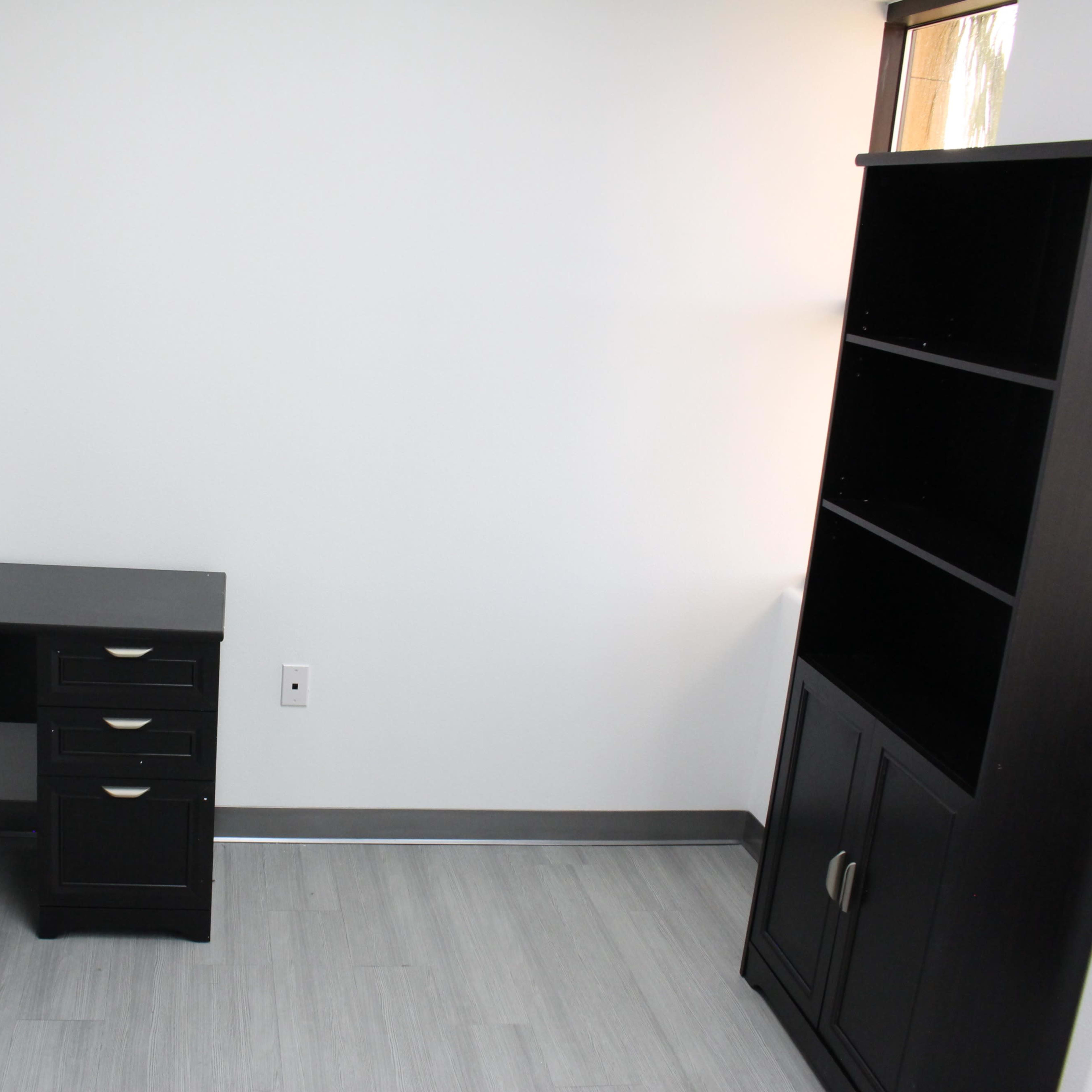 An empty office space featuring a black desk with drawers and a black office chair, alongside a tall shelf unit against a white wall.
