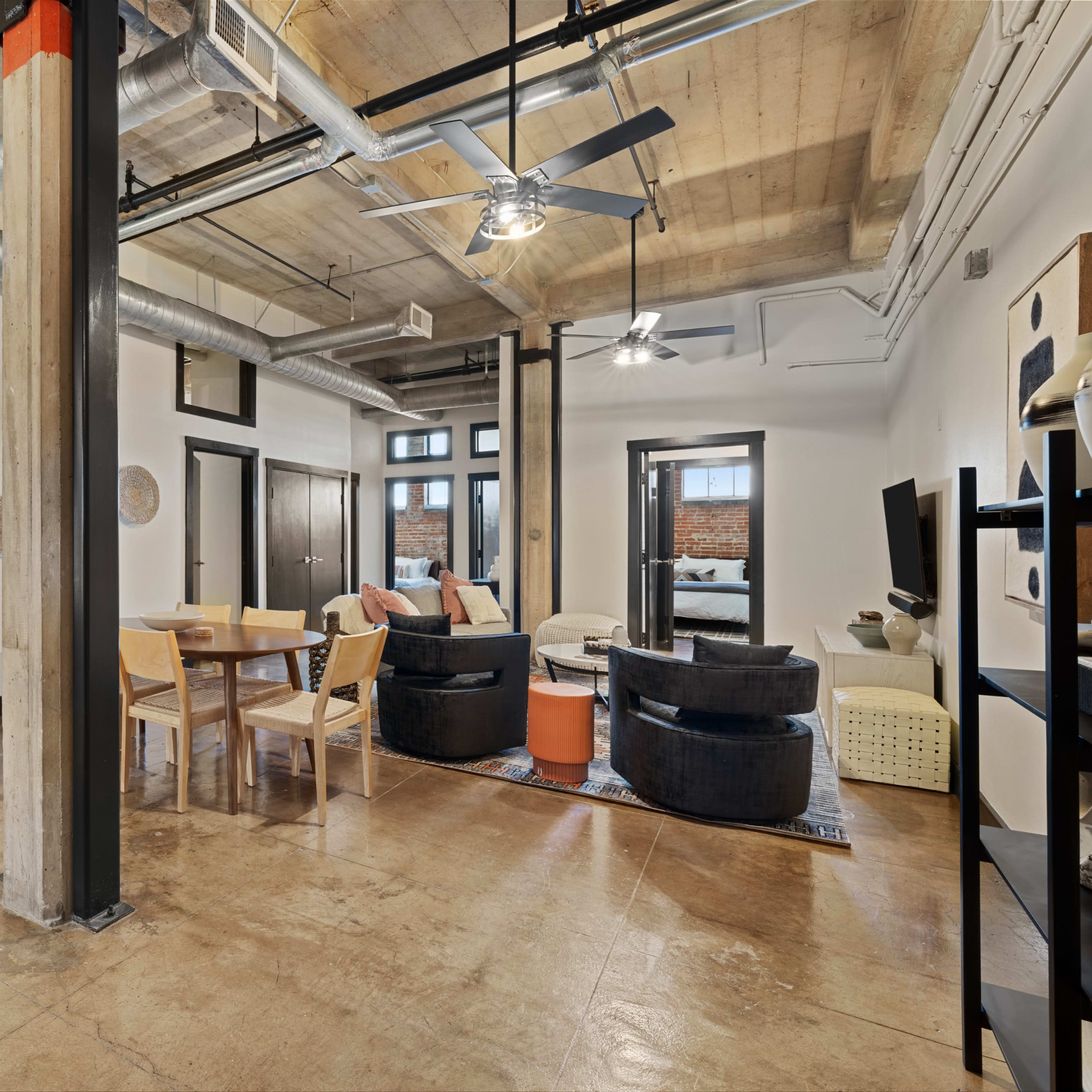 A modern loft-style living space featuring an open layout with a dining area, seating arrangement, and industrial-style elements such as exposed beams and ductwork.
