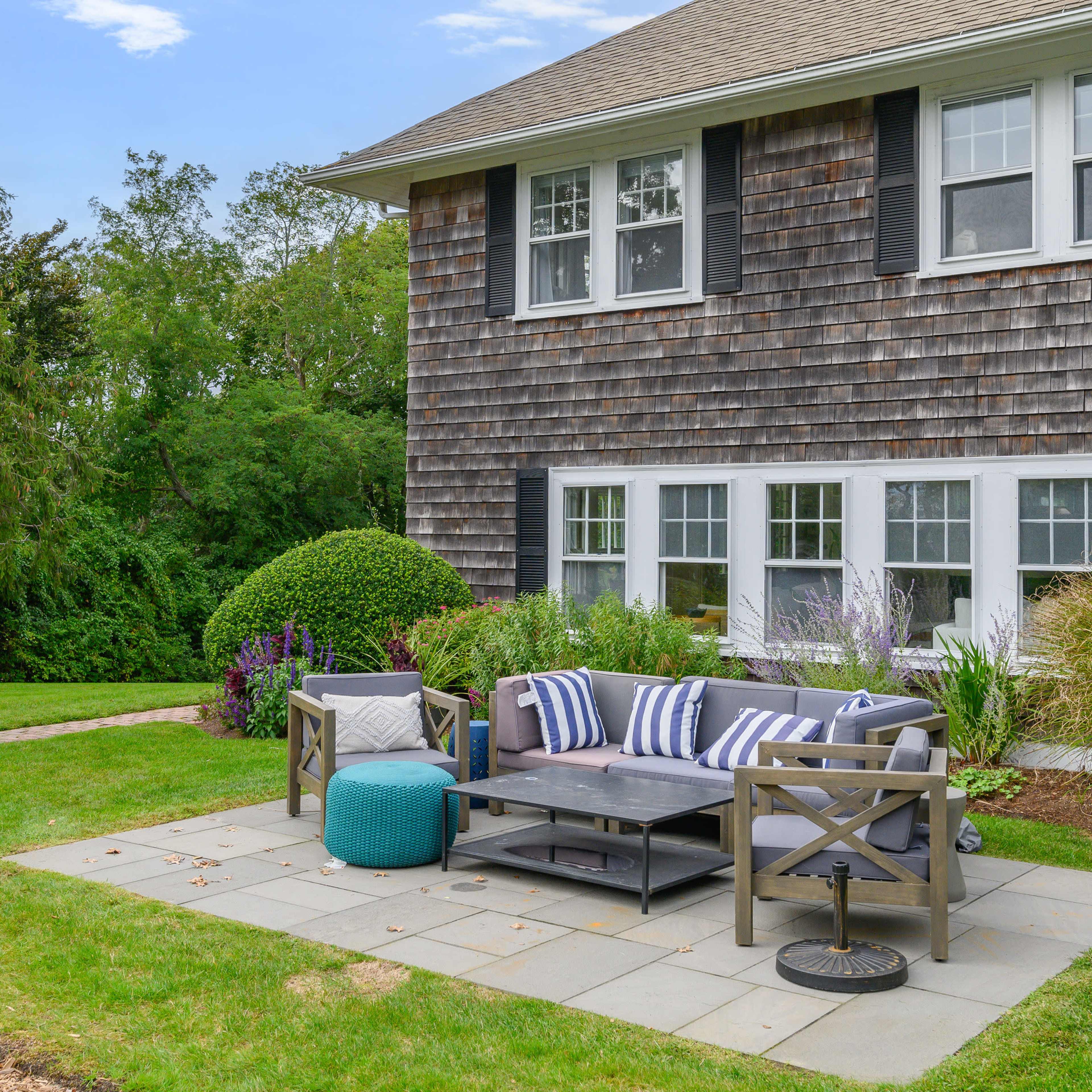 A patio area with a gray couch, blue ottoman, and coffee table situated beside a house with wooden shingles and large windows.