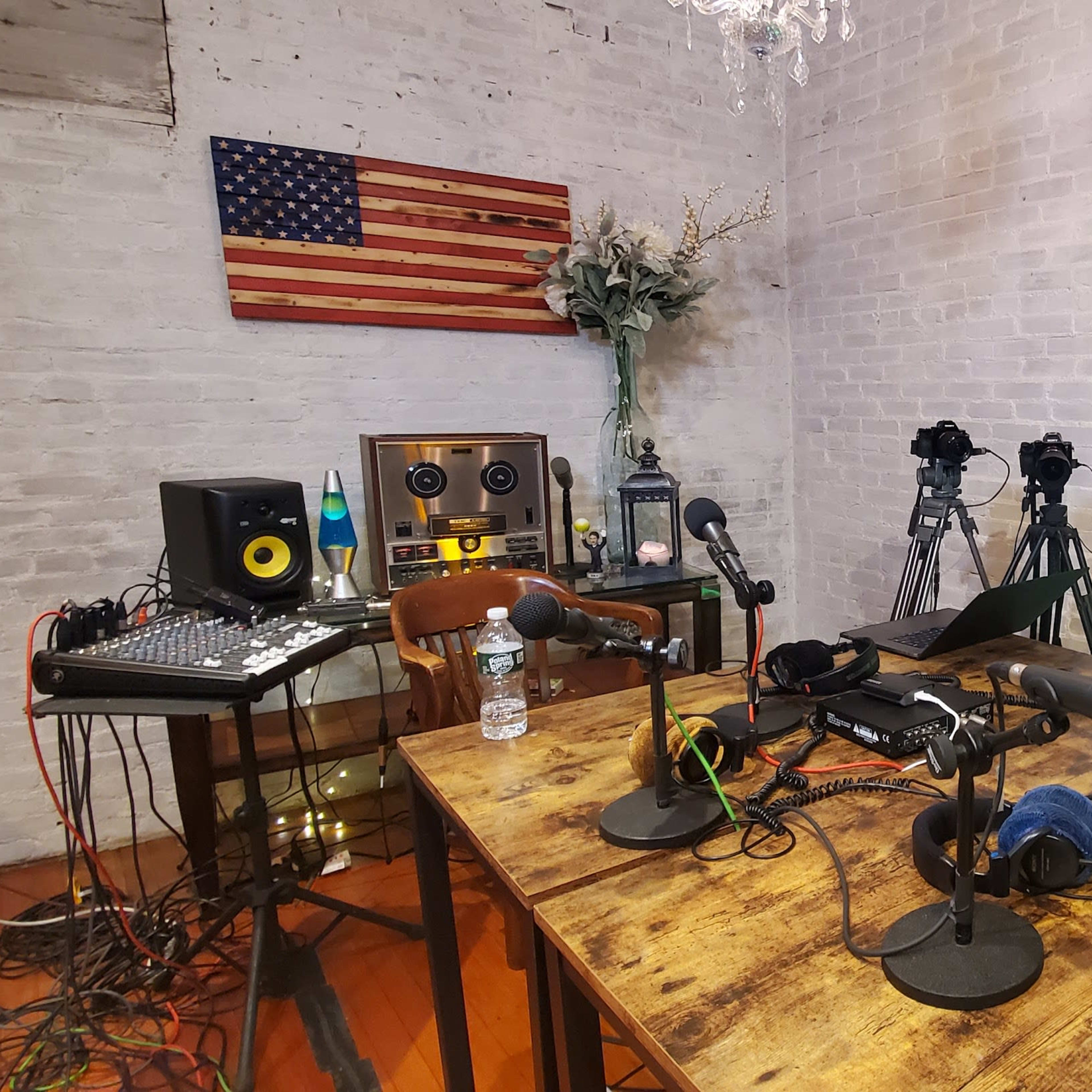 A recording studio setup featuring multiple microphones, cameras, and audio equipment against a backdrop of a wooden flag and a potted plant.