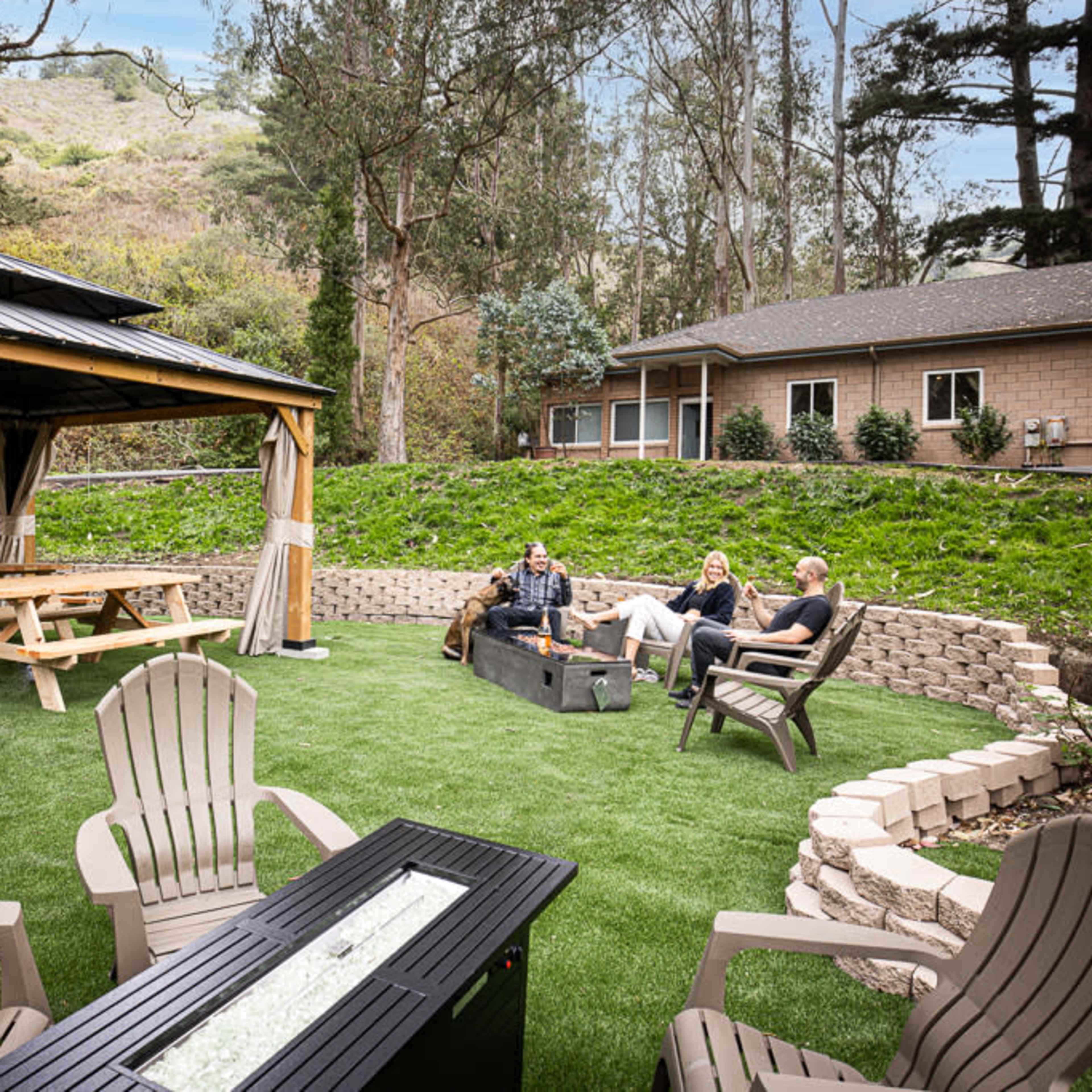 A group of three people sits around a fire pit on a landscaped lawn near a gazebo and a building surrounded by trees.