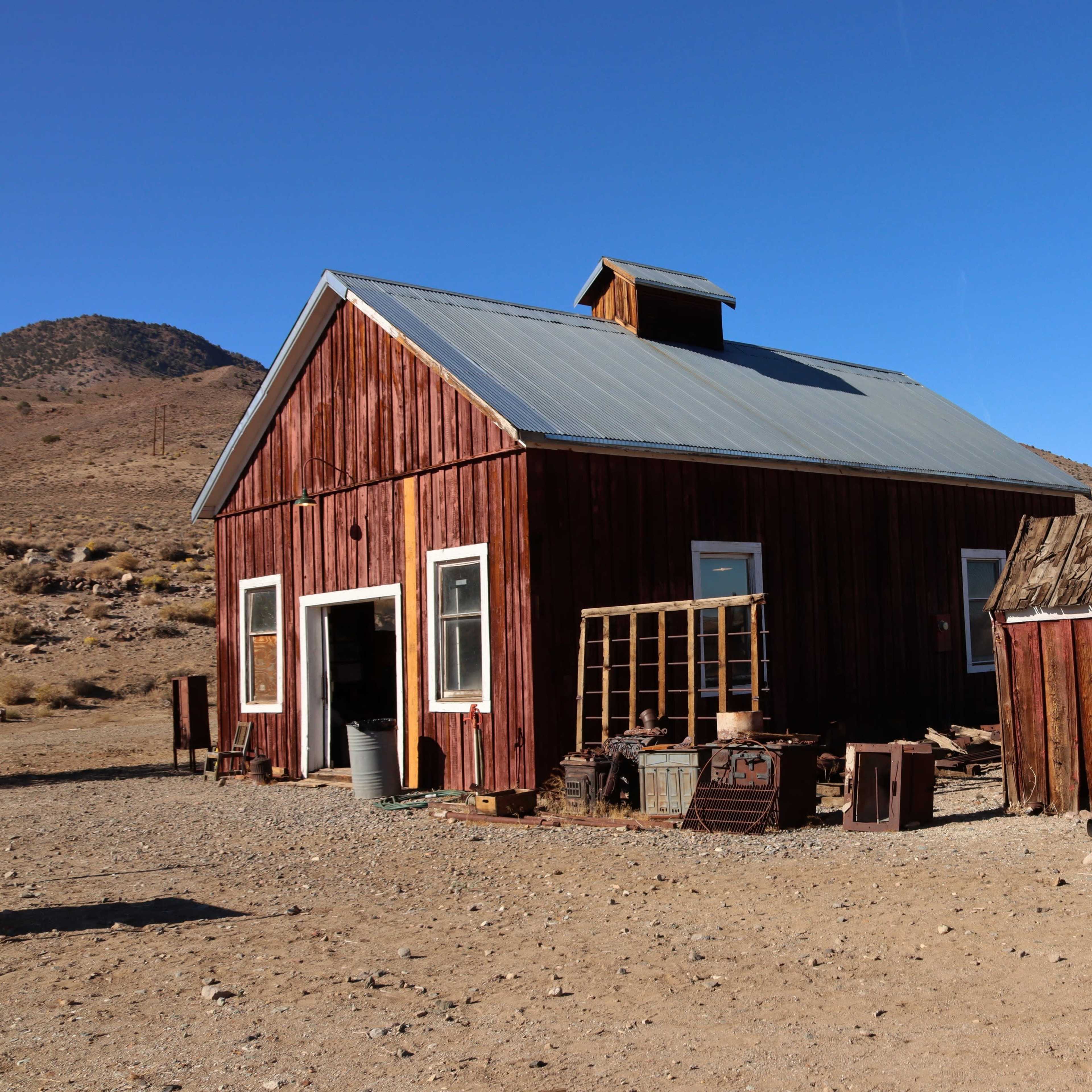 A red wooden house with a metal roof stands next to a smaller, weathered shed in a dry, desert landscape.